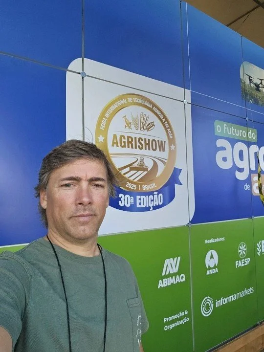 A man taking a selfie in front of a large blue and green banner at the AGRISHOW agricultural technology fair in Brazil, 2025.
