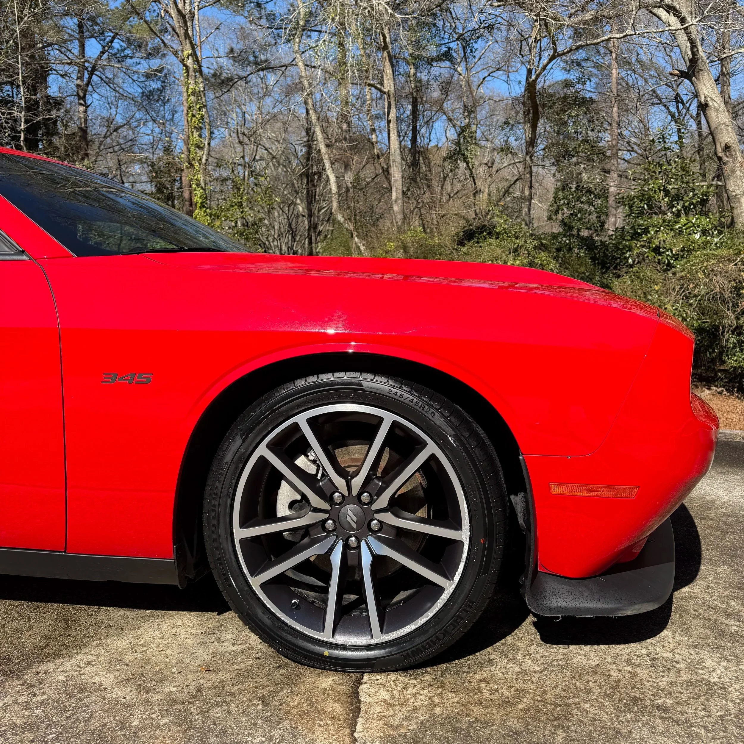 Close-up of the front left side of a red sports car with a black tire, alloy rim, and the badge '345' on the side, parked on pavement with trees in the background.