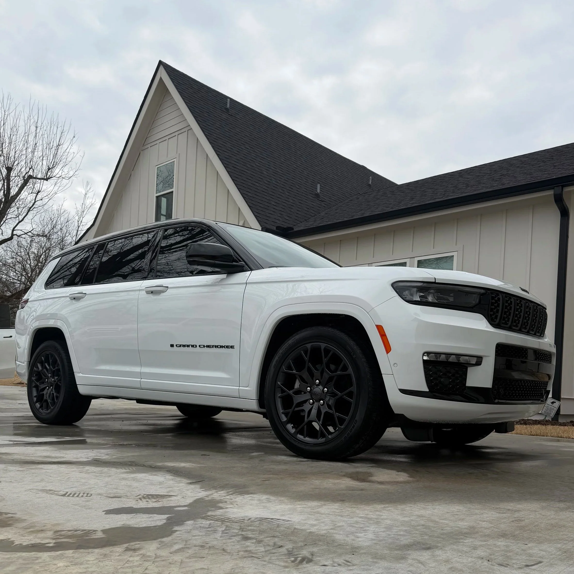 White Jeep Grand Cherokee parked on driveway in front of a modern house with black roof shingles and white siding.