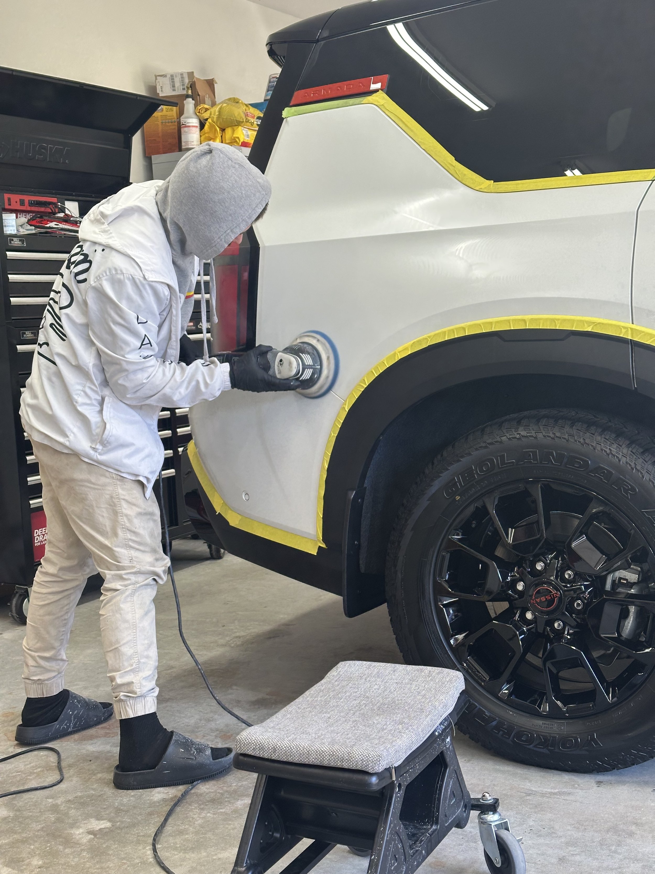 A person wearing a hoodie and gloves is polishing a white vehicle with a power buffer inside a garage.