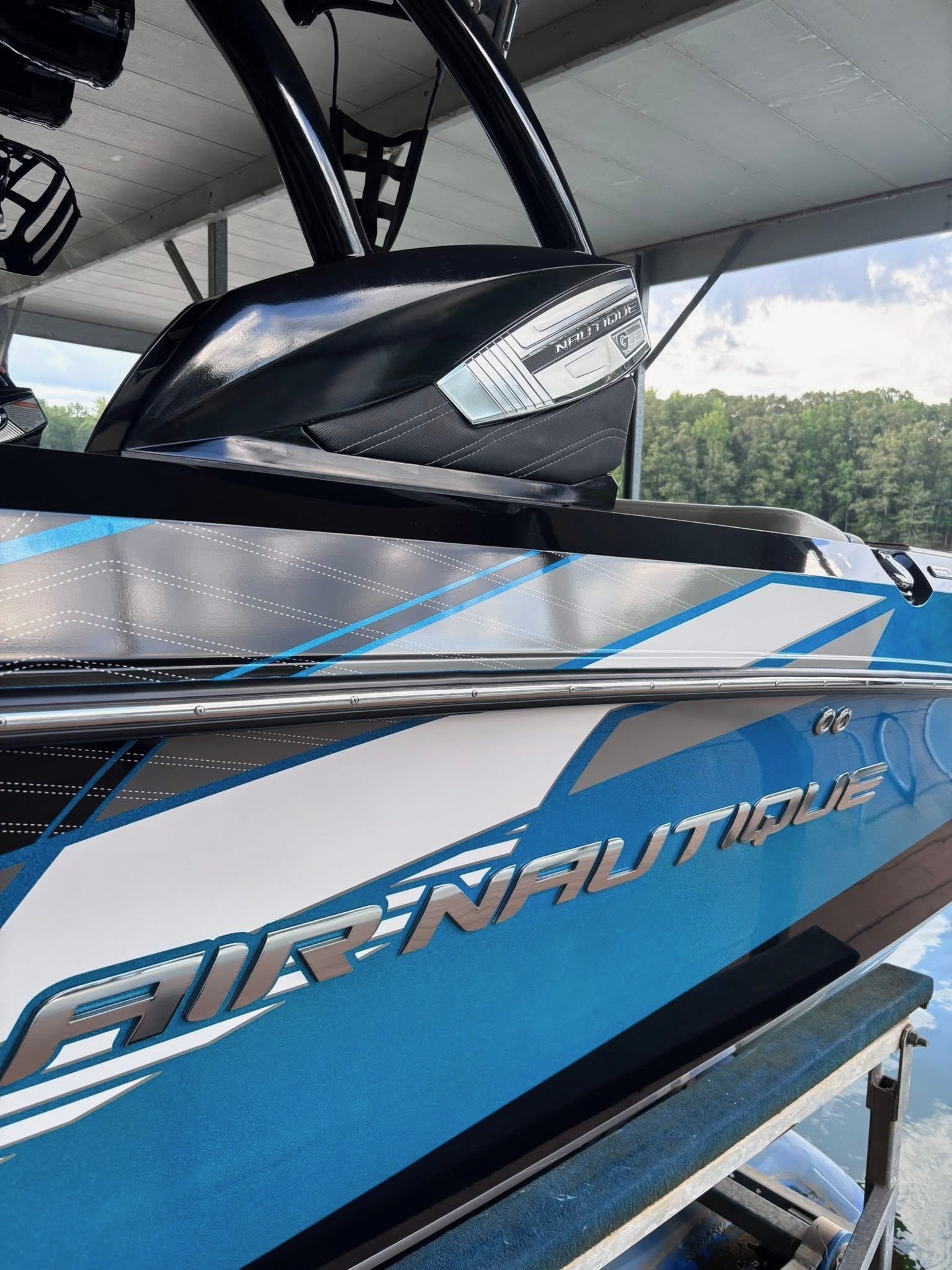 Blue and black Nautique wakeboarding boat on a boat lift under a roof, with a green wooded landscape in the background.