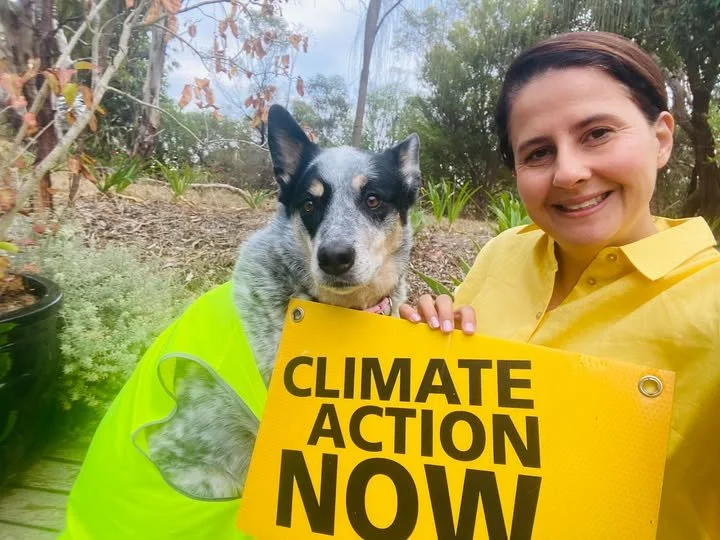 A woman in a yellow shirt holding a sign that says 'CLIMATE ACTION NOW' next to a dog in a green vest outside in a garden.