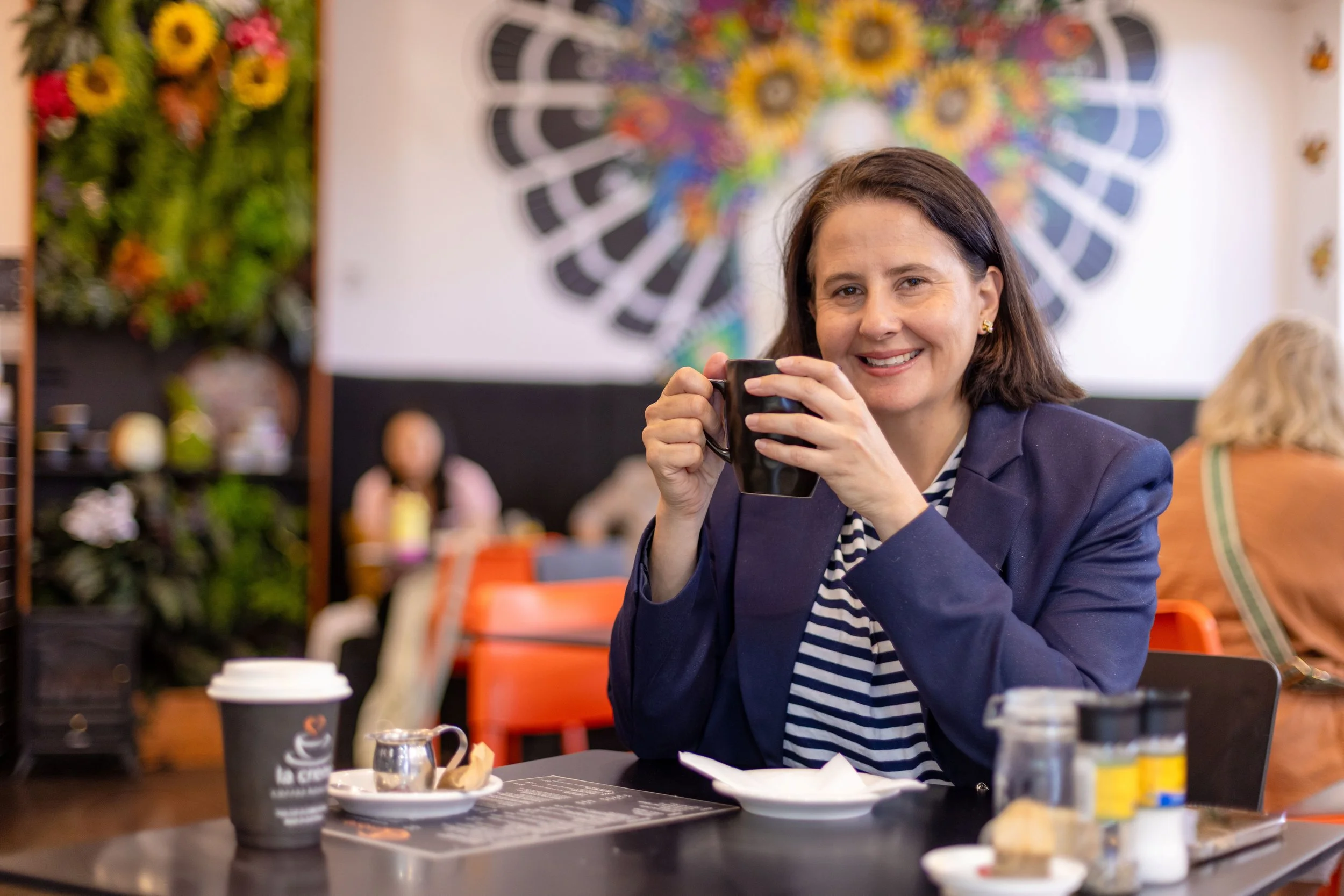 A woman with dark hair, wearing a navy blazer and striped shirt, sitting at a table in a colorful, decorated cafe, holding a black mug and smiling at the camera. The table has a paper coffee cup, small pitcher, and some food items.