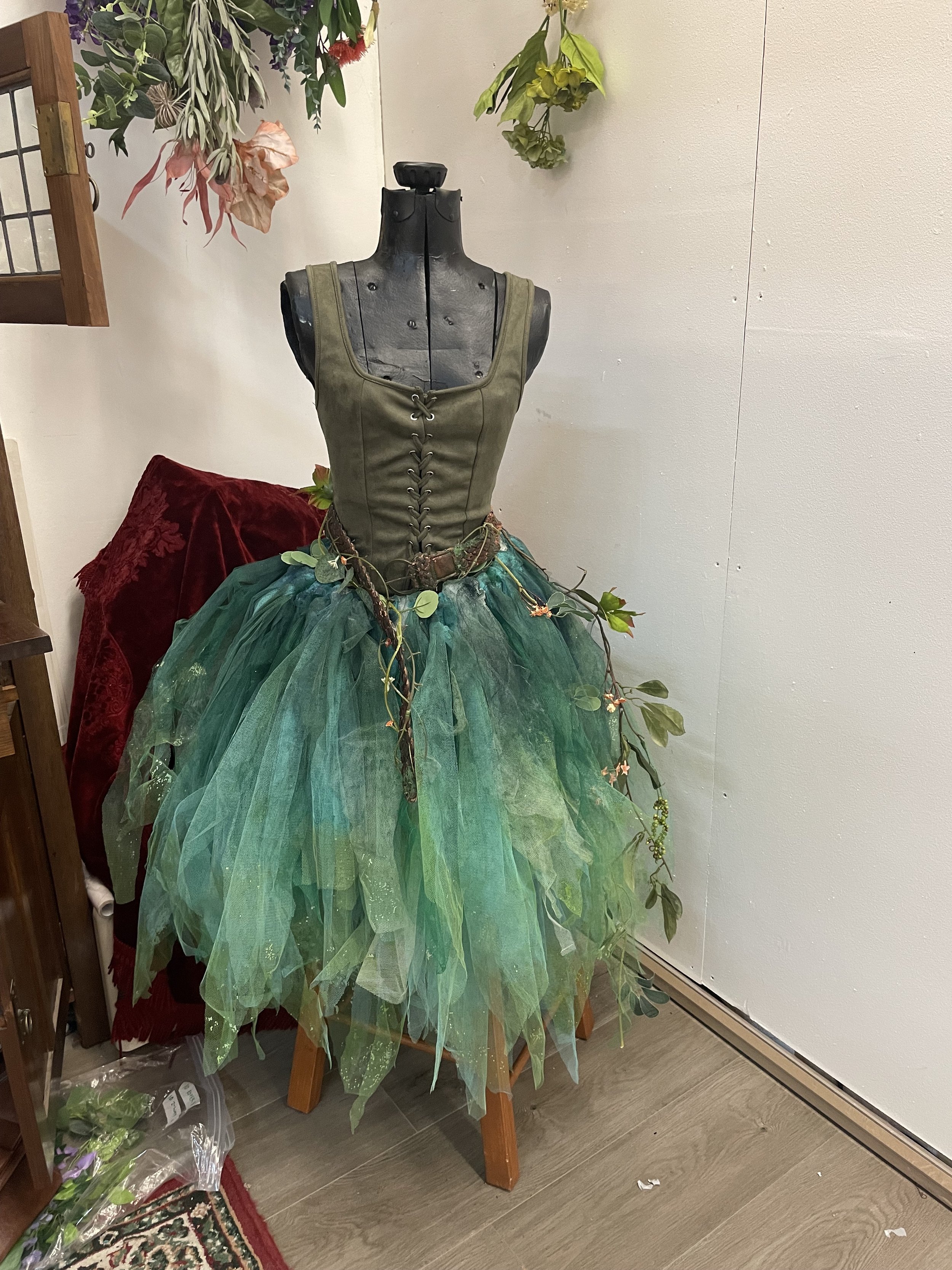A mannequin torso dressed in a green dress with a lace-up bodice, paired with a green tulle skirt decorated with vines and flowers, displayed indoors near a white wall, with some flowers and other display items around.