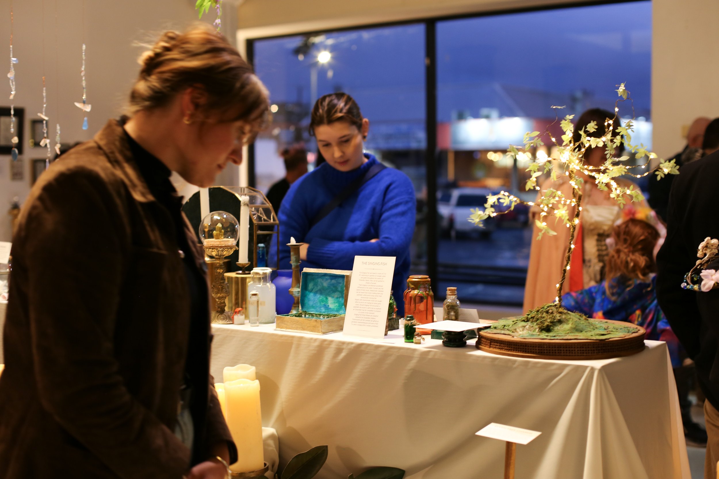 People browsing art and craft displays at an indoor market or exhibition during evening hours.
