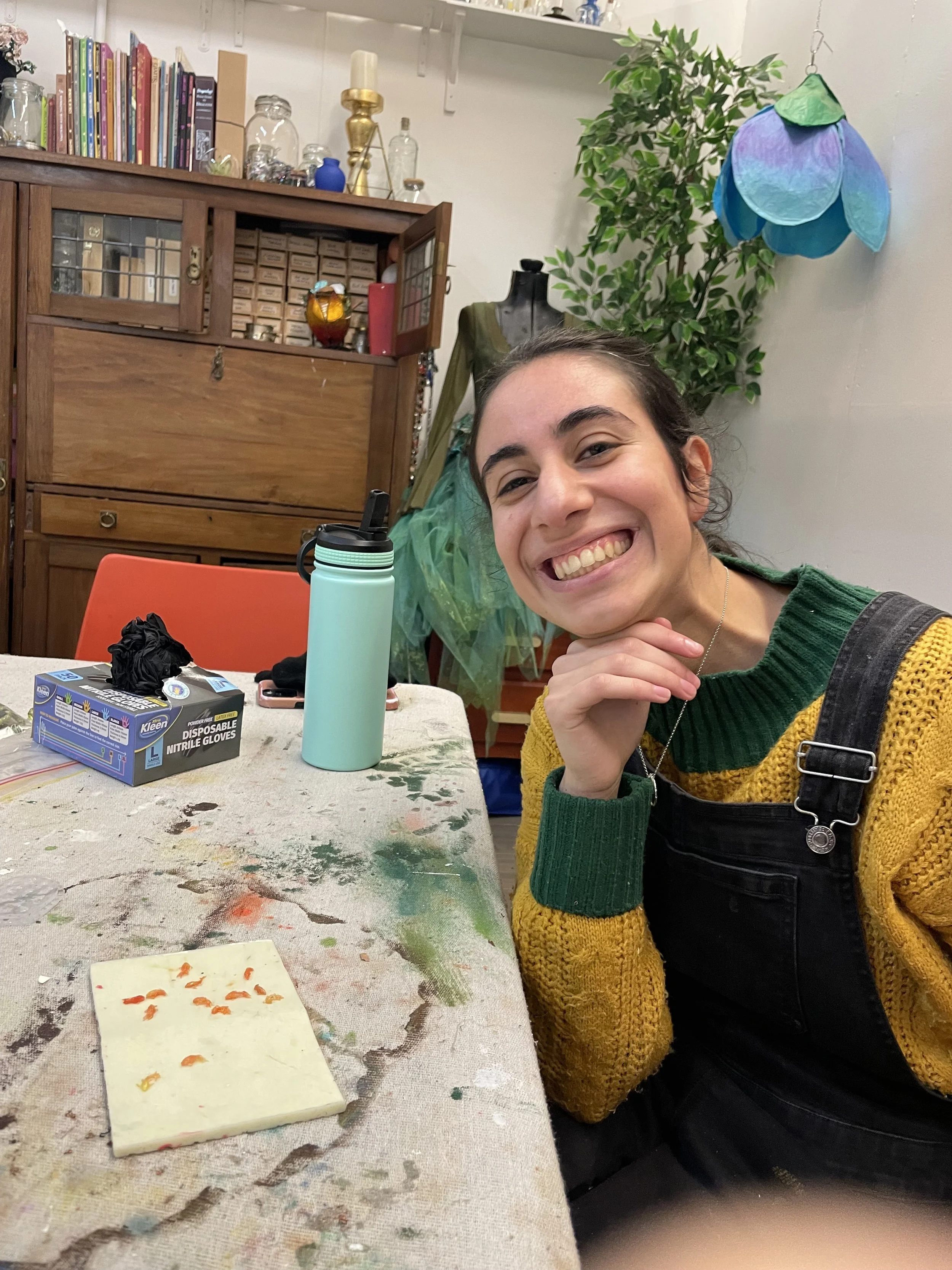A young woman smiling while sitting at a table with paint smudges, with small orange paint pieces on a paper in front of her. The background shows a bookshelf, a mannequin with a green garment, plants, and hanging lamps in an art or craft studio.