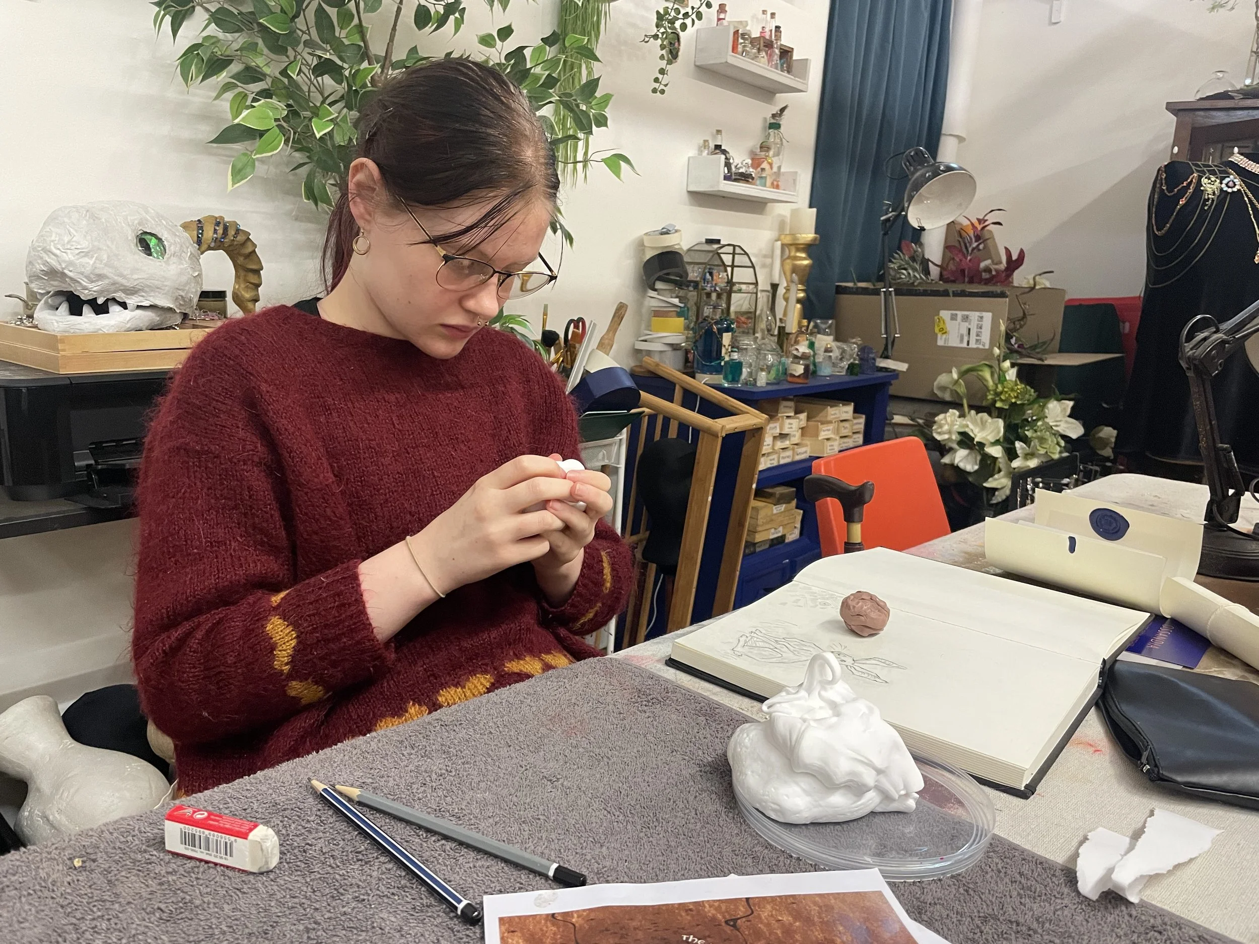 A woman with glasses and a septum piercing is working on an art project at a cluttered table. She is wearing a red sweater with yellow and orange pattern details. There is a large, realistic skull model and a clay sculpture of a head on the table. Th