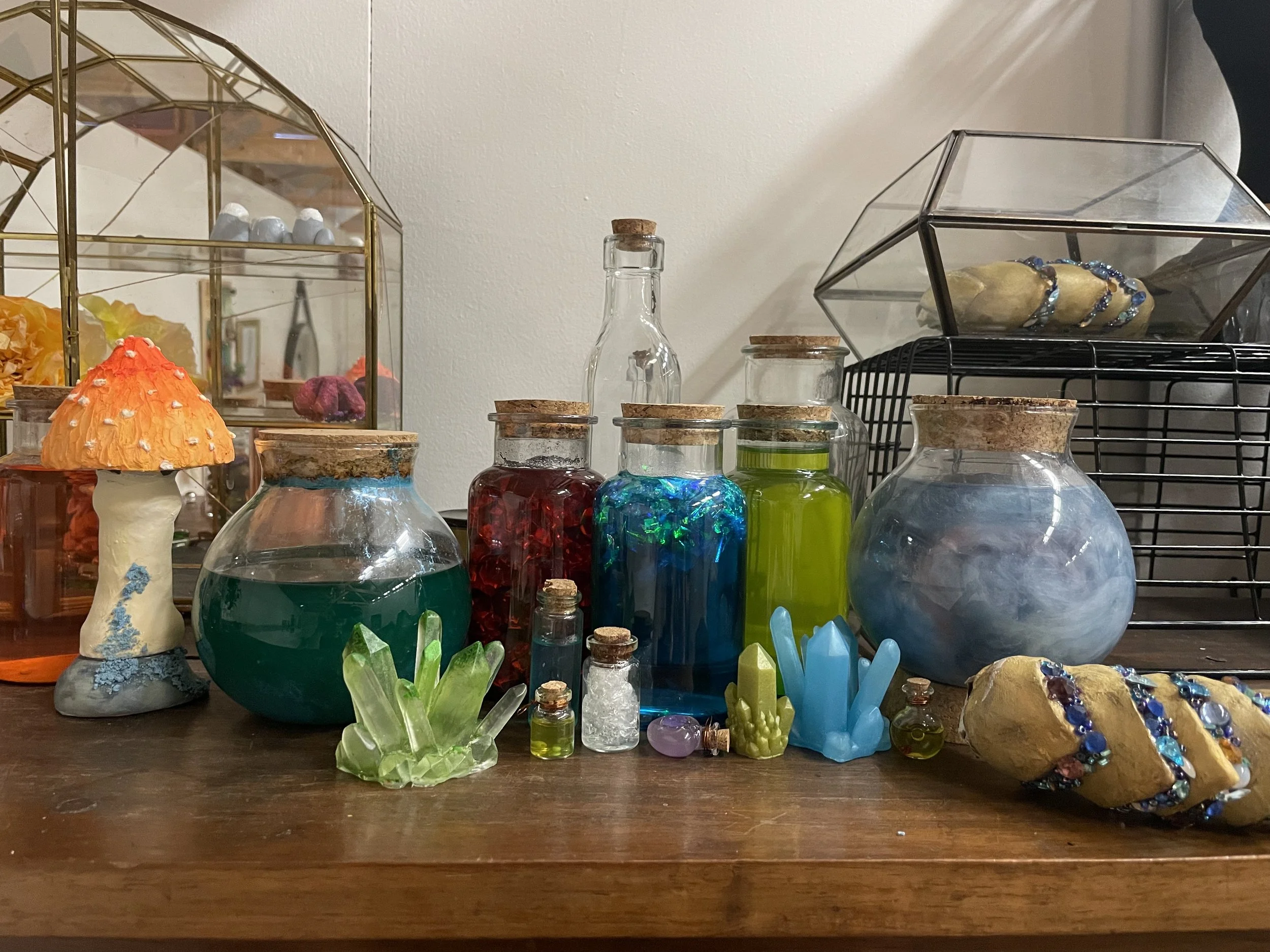 A collection of colorful glass bottles and jars with cork stoppers, decorative mushrooms, crystals, and jewelry displayed on a wooden surface against a plain wall.