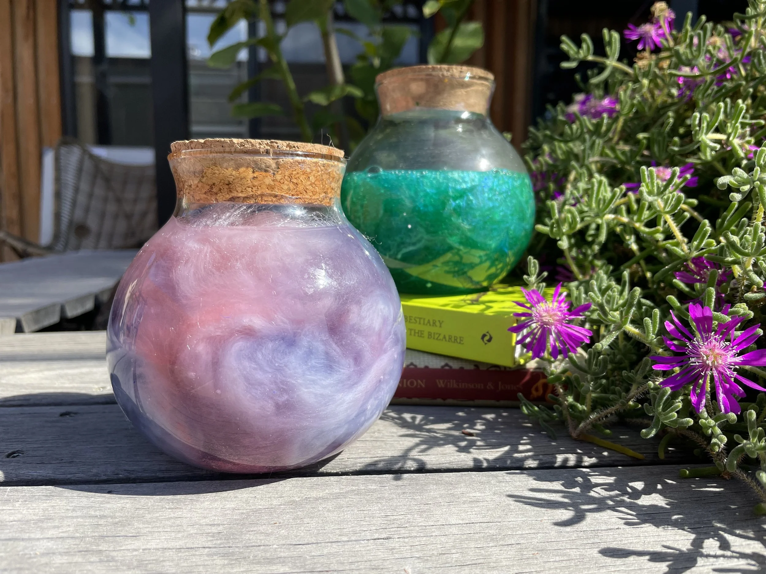 Two small glass vases with cork stoppers, one pink and one green, placed on a wooden surface outdoors with purple flowering plants nearby.
