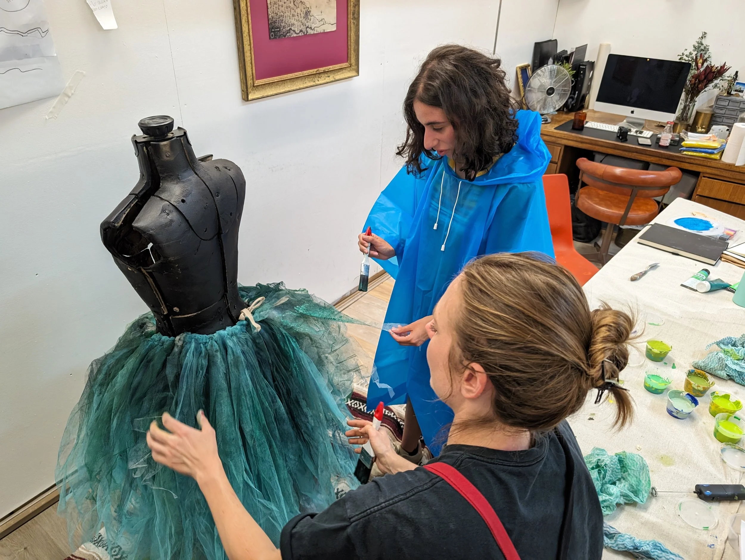 People painting a dress on a mannequin in an indoor art studio with paints and brushes on a table.