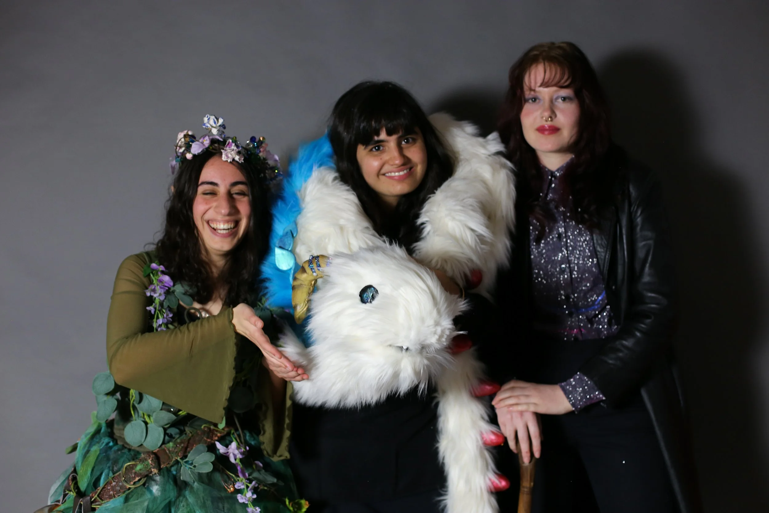 Three women standing together, smiling, with a large white fluffy dog. The woman on the left has long dark hair, a flower crown, and a green outfit adorned with flowers. The woman in the middle has short dark hair and is wearing a white fur cape. The
