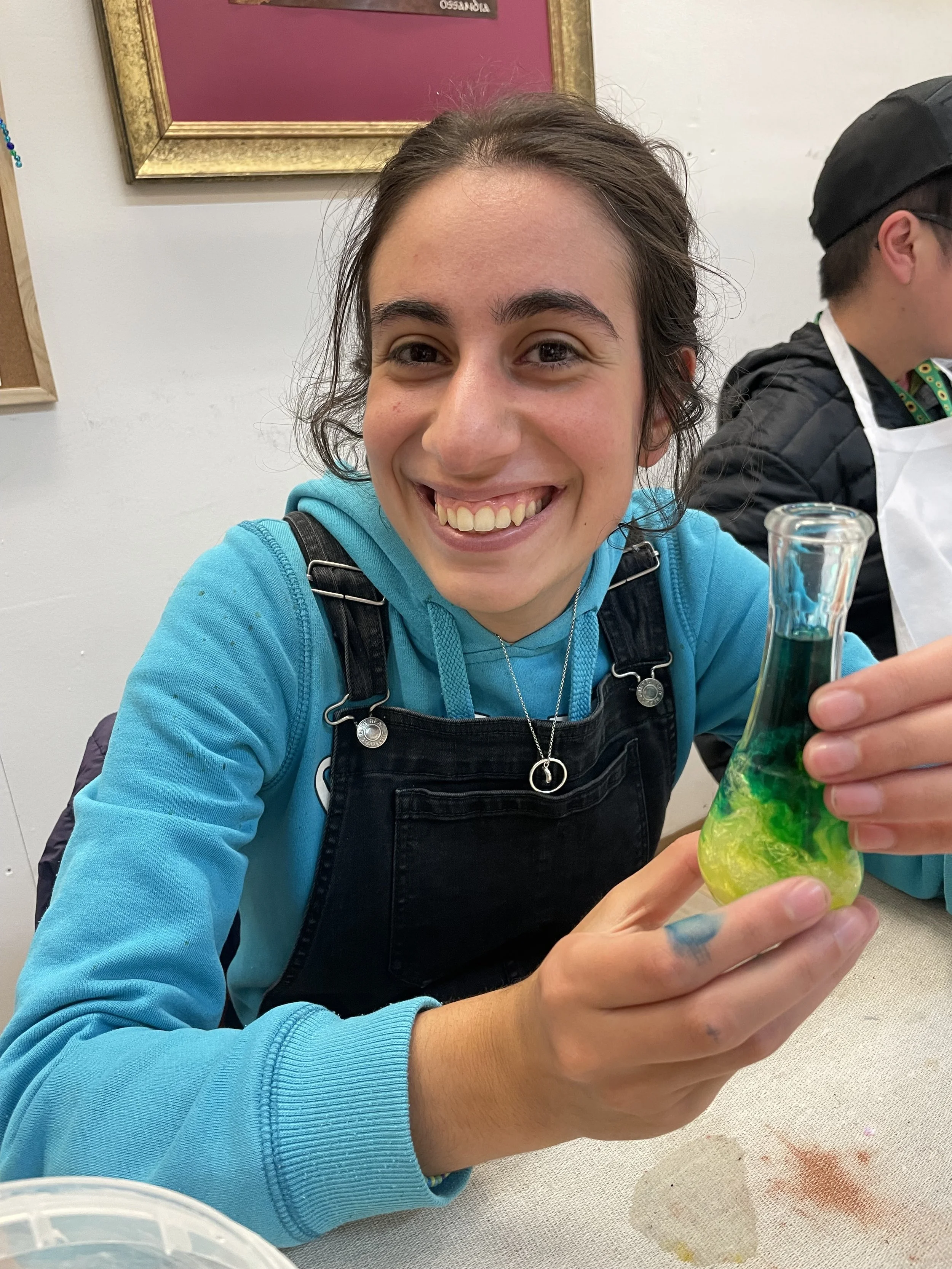 A young woman smiling and holding a colorful marble or glass art piece in her hand, sitting at a table with art supplies. She is wearing a blue hoodie and a black apron, and there is a cluttered art workspace behind her.