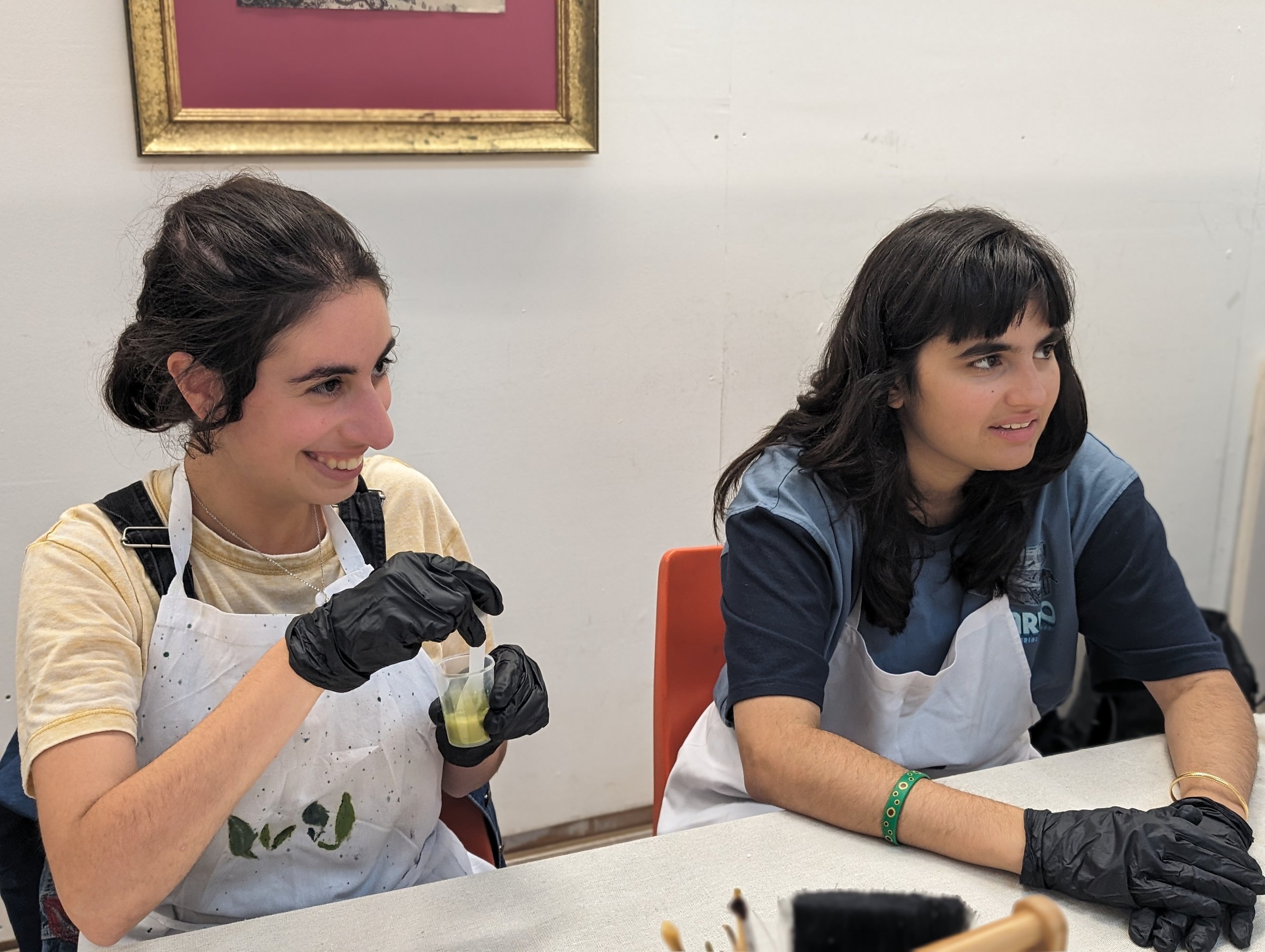 Two young women sit at a table, engaged in an activity. The woman on the left has dark hair tied back, is smiling, and wearing a yellow shirt under a black apron, holding a cup of green juice and wearing black gloves. The woman on the right has dark 