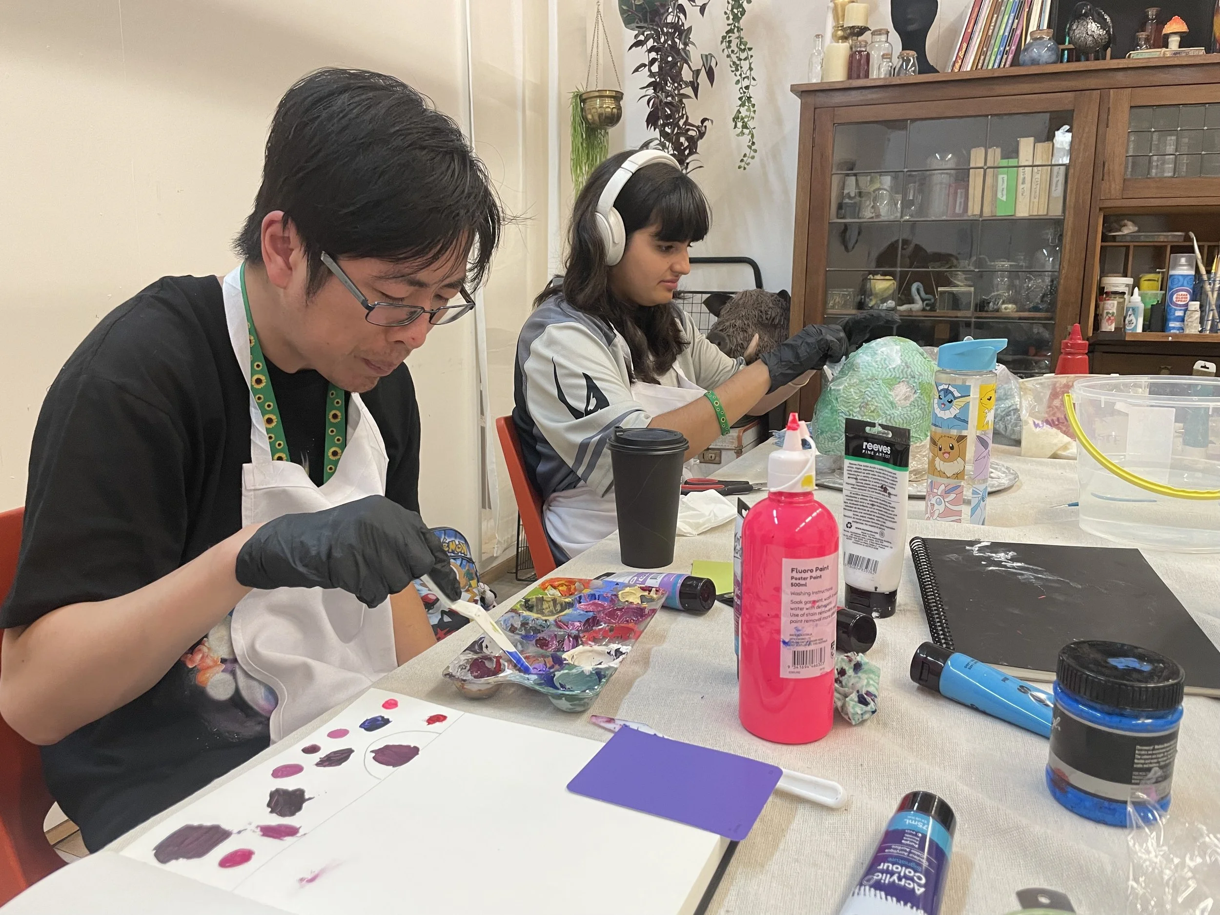 People participating in a painting activity at a table with art supplies, including paints, brushes, and bottles, in a room decorated with plants and shelves filled with books and decorative items.
