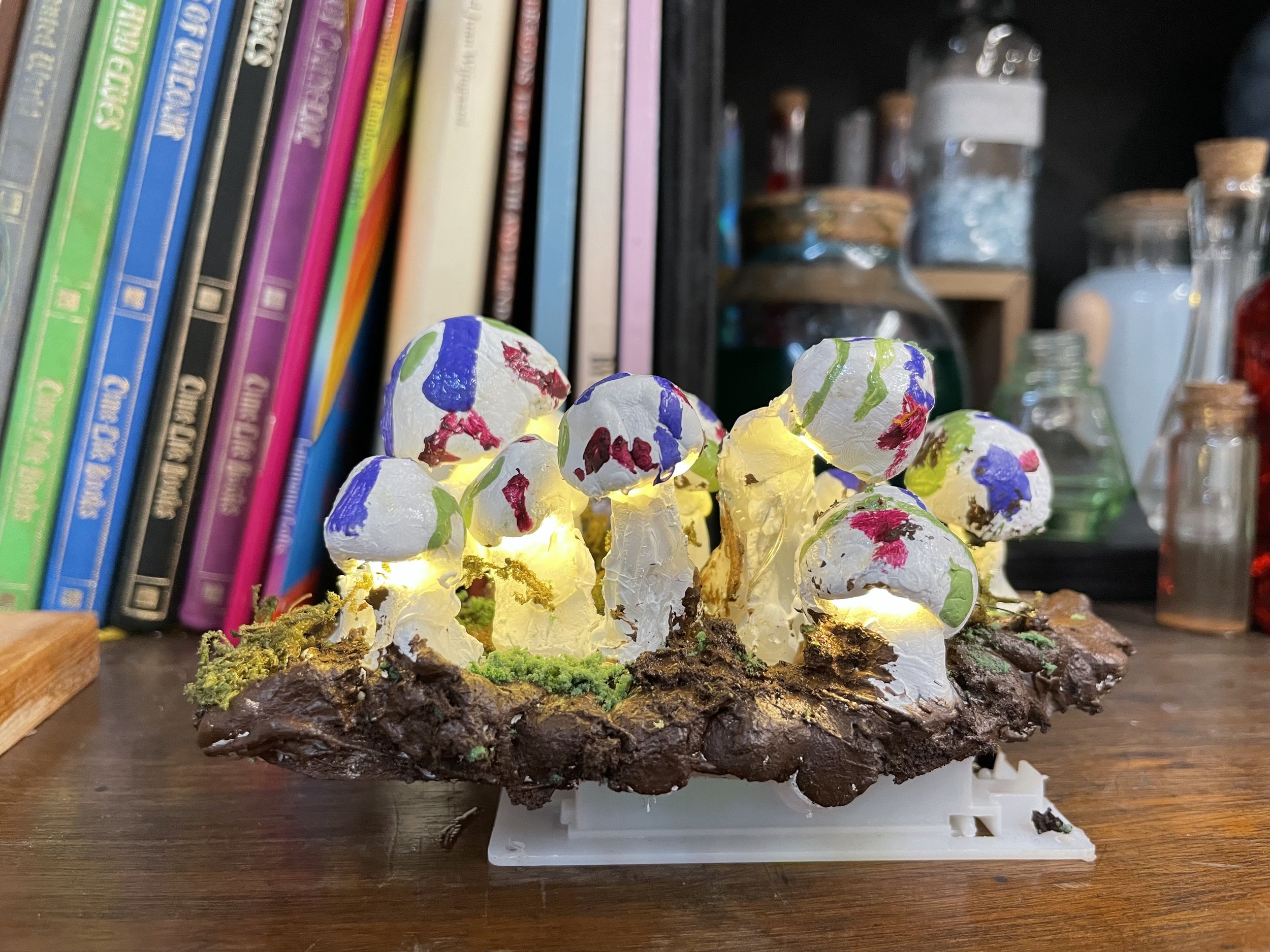 Miniature model of a light up mushrooms with colorful painted designs, set on a wooden surface, with books and laboratory glassware in the background.