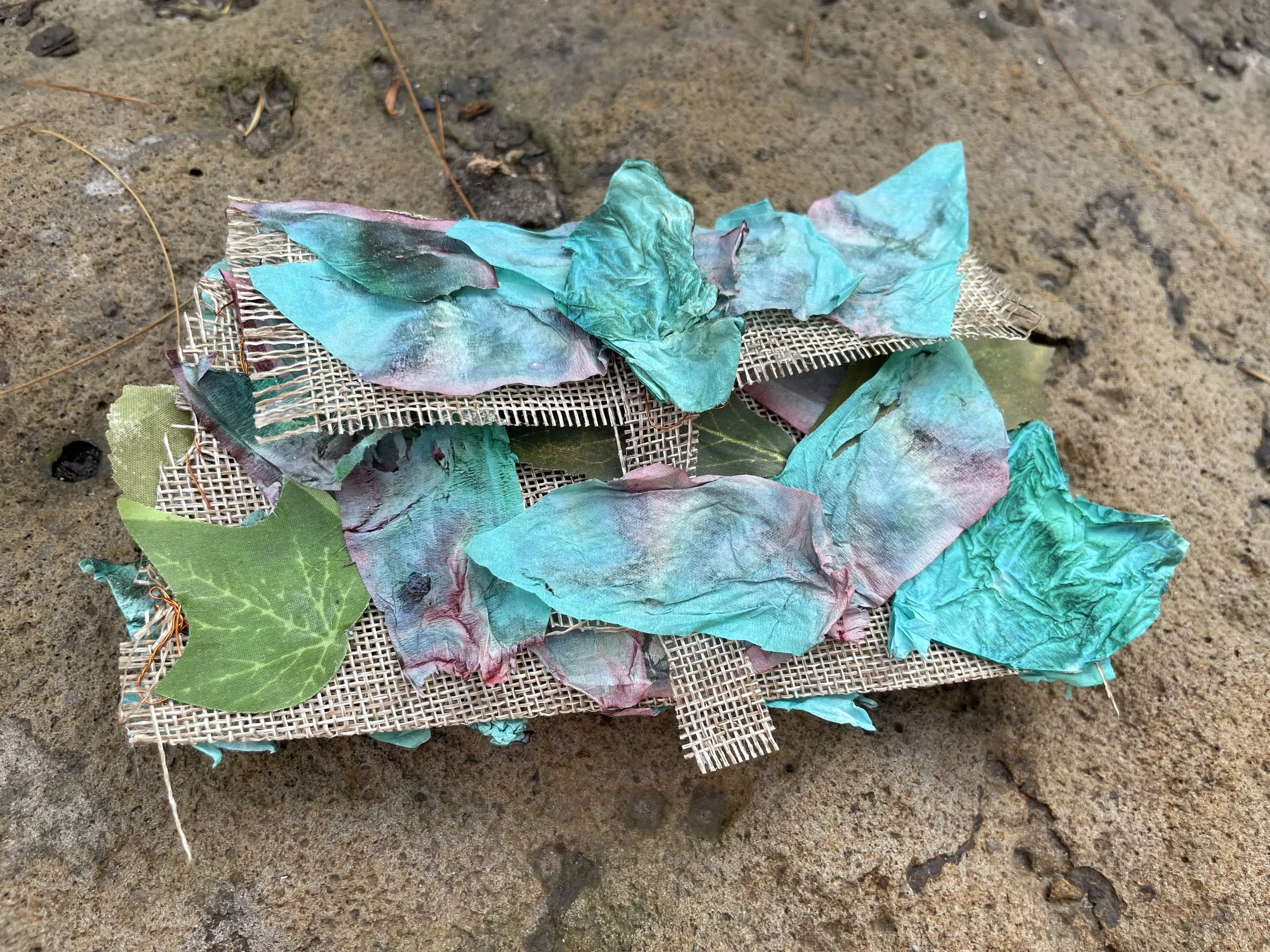 Colorful crumpled tissue papers scattered on plastic mesh, situated on sandy ground with small rocks and twigs around.