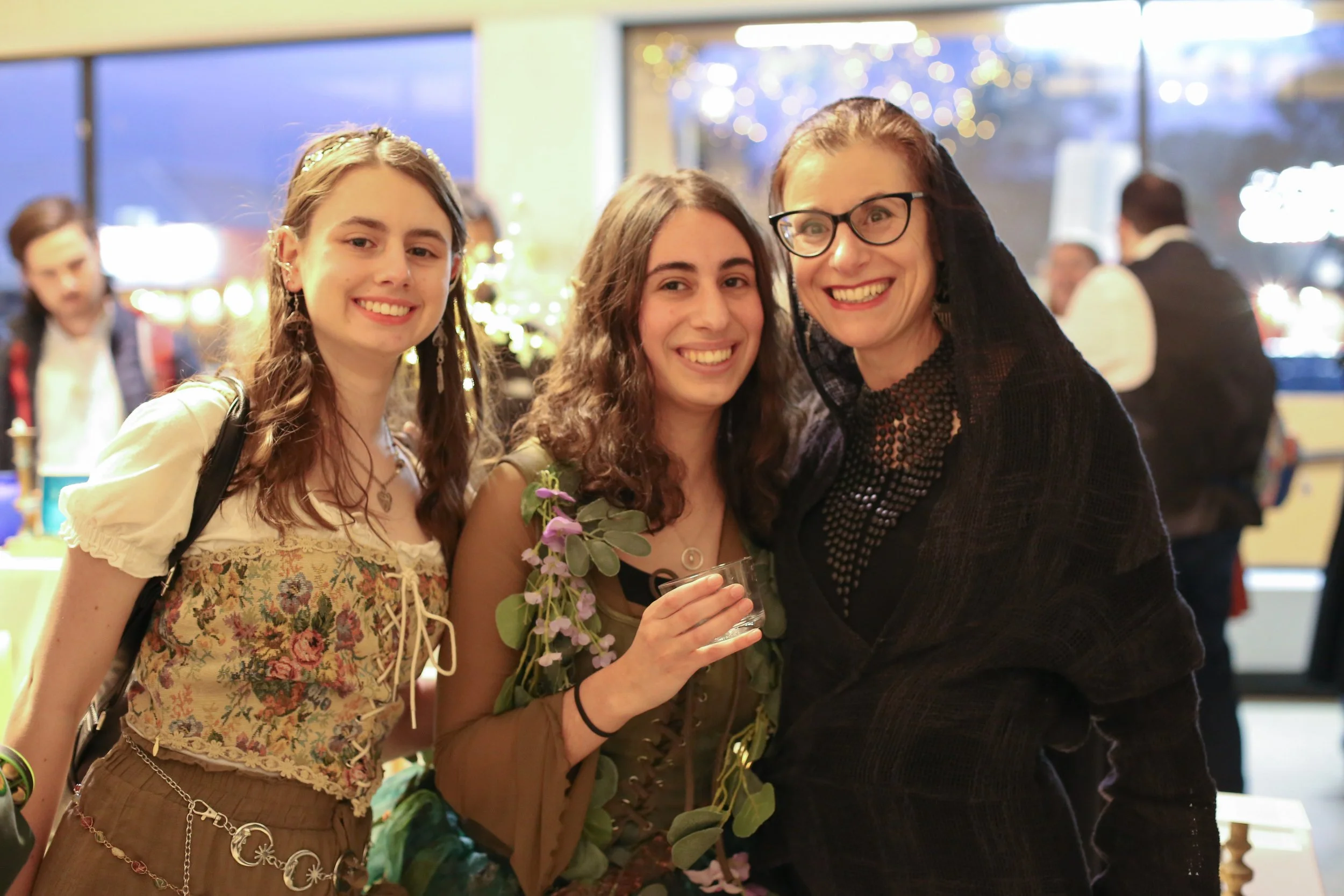Three women smiling at a social gathering, with two women in the foreground and one in the background, all wearing festive or vintage-style clothing.