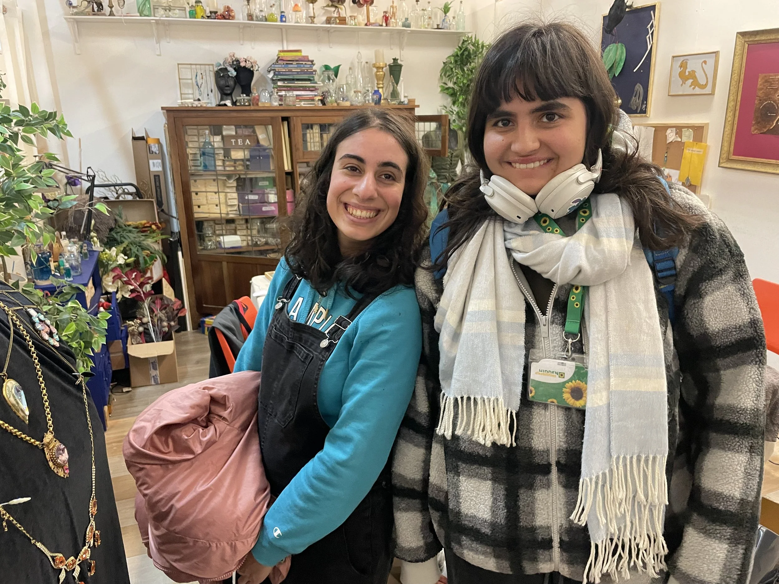 Two women smiling in a store with shelves filled with decorative bottles, books, and artwork in the background. One woman has curly dark hair, and the other has wavy dark hair and is wearing headphones around her neck.