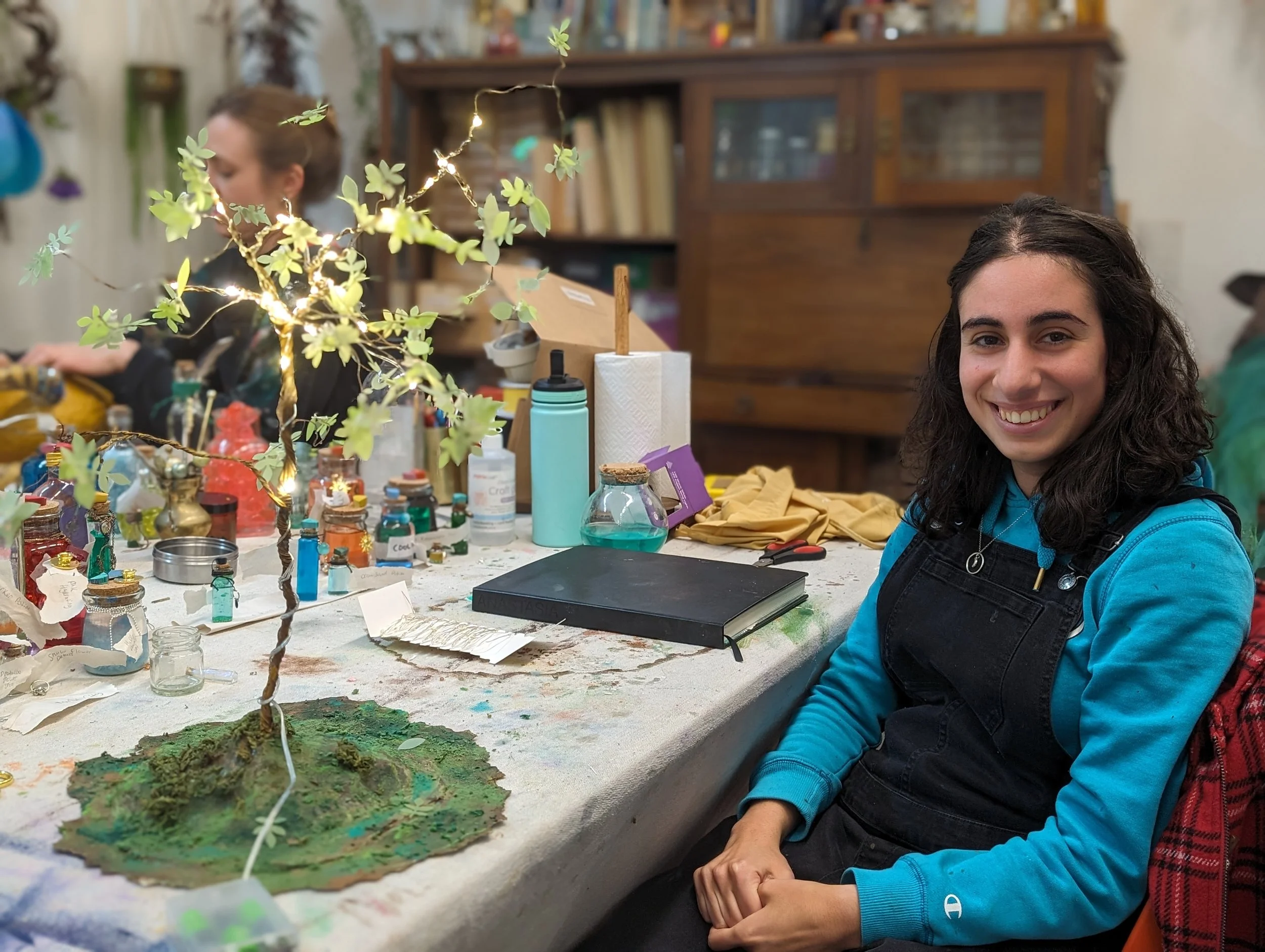A young woman with curly hair smiling at the camera, sitting at a cluttered table with art supplies, a small artificial tree with lights and leaves, and a miniature landscape; another person is blurred in the background in an art or craft studio.