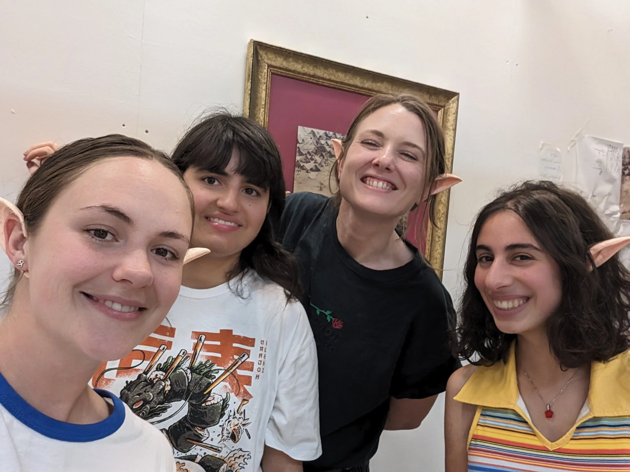Group of young women dressed as elves with pointed ears, smiling indoors in front of a framed picture on the wall.