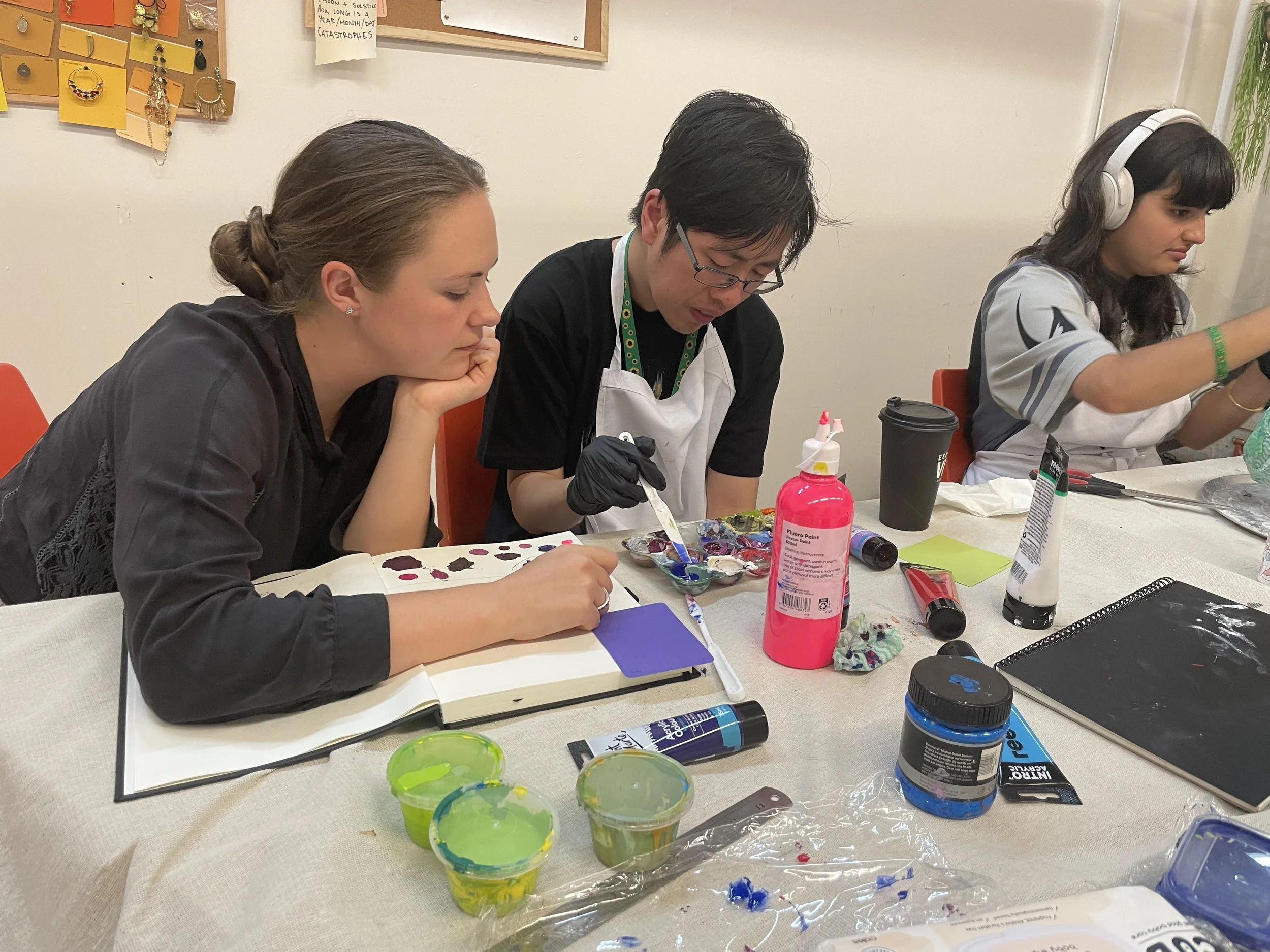 Three people seated at a table engaged in a painting activity. There are various art supplies including paint palettes, brushes, and cups of paint in front of them. The background features a bulletin board and jewelry hanging on the wall.