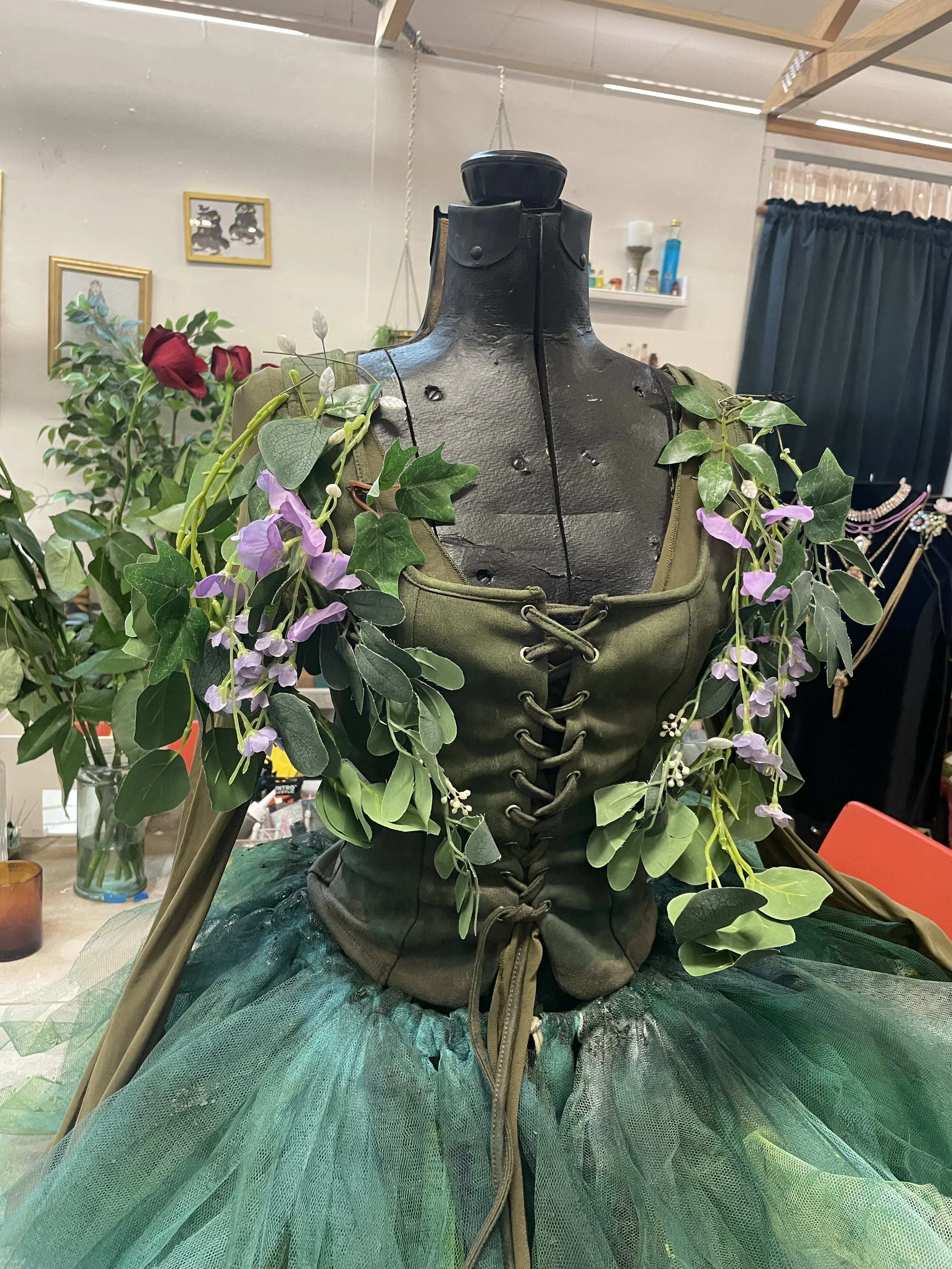 A costume dress form with artificial green leaves, purple flowers, and some red roses draped over the shoulders. The dress form is in front of a room with shelves, framed pictures, and various bottles and items.