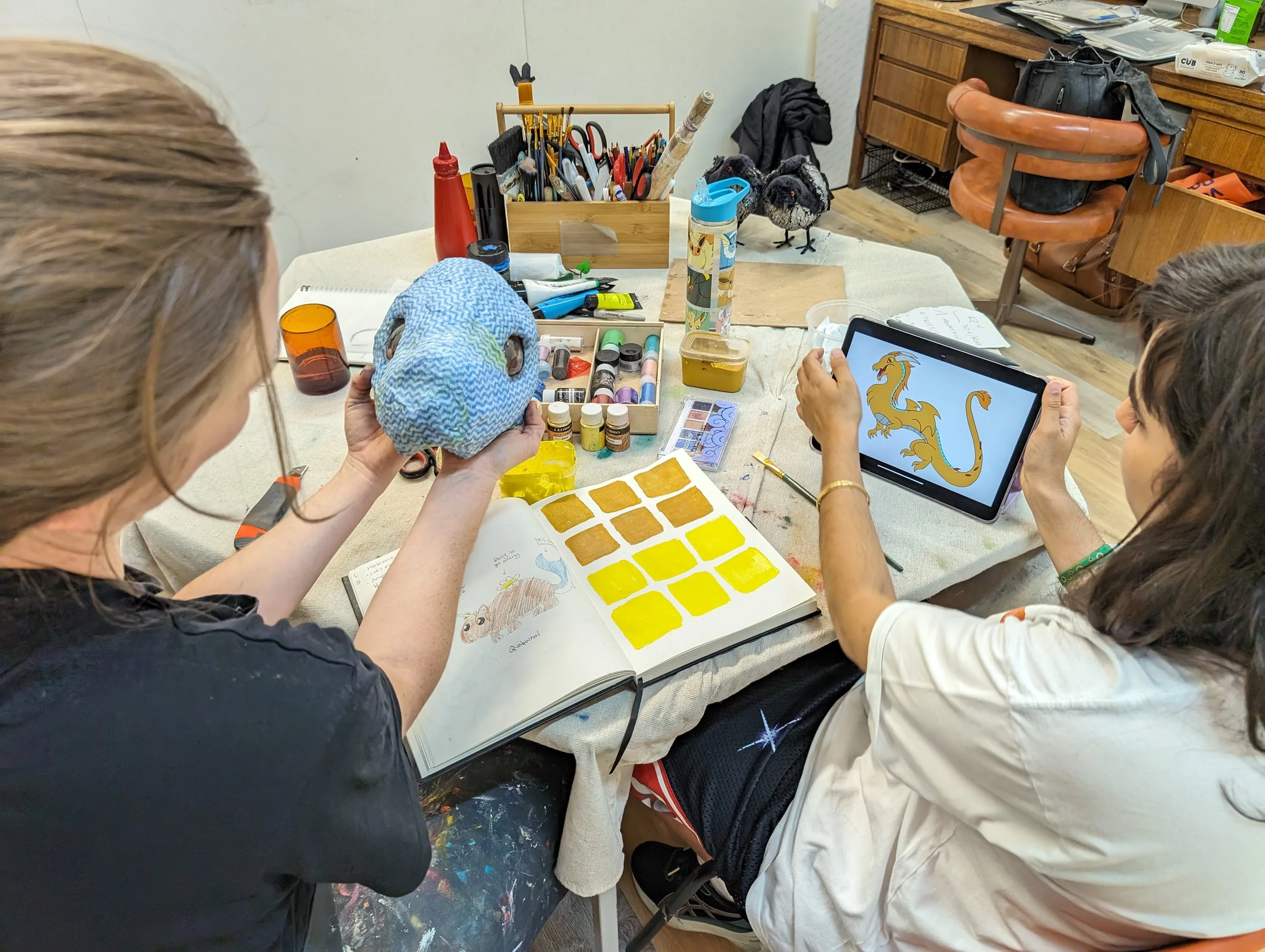 People gathered around a cluttered table with art supplies, an open sketchbook, and a tablet displaying a drawing of a dragon.