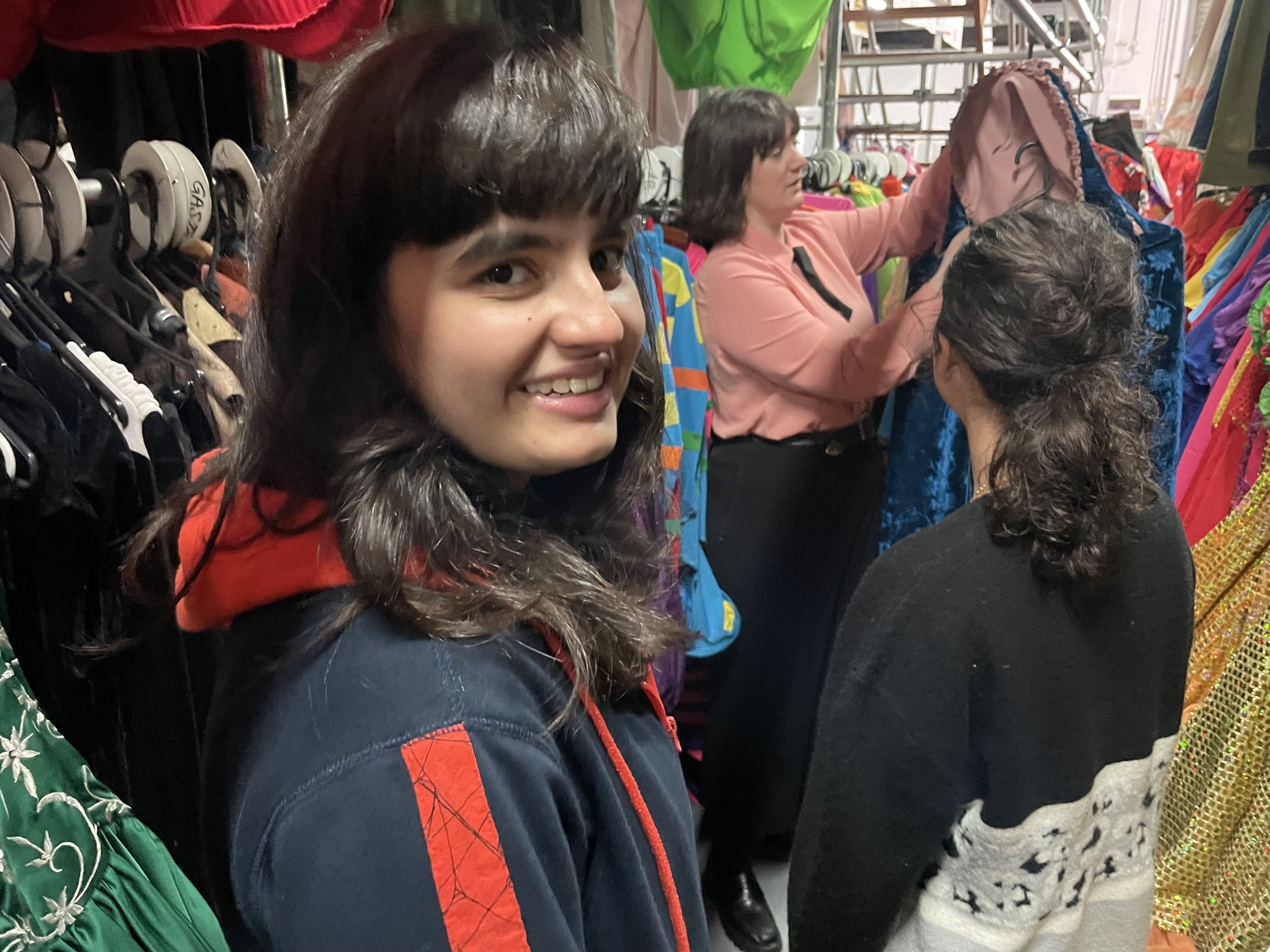 A young woman with dark brown hair and a smile, wearing a dark jacket with red accents, shopping among racks of colorful clothing and accessories in a store.