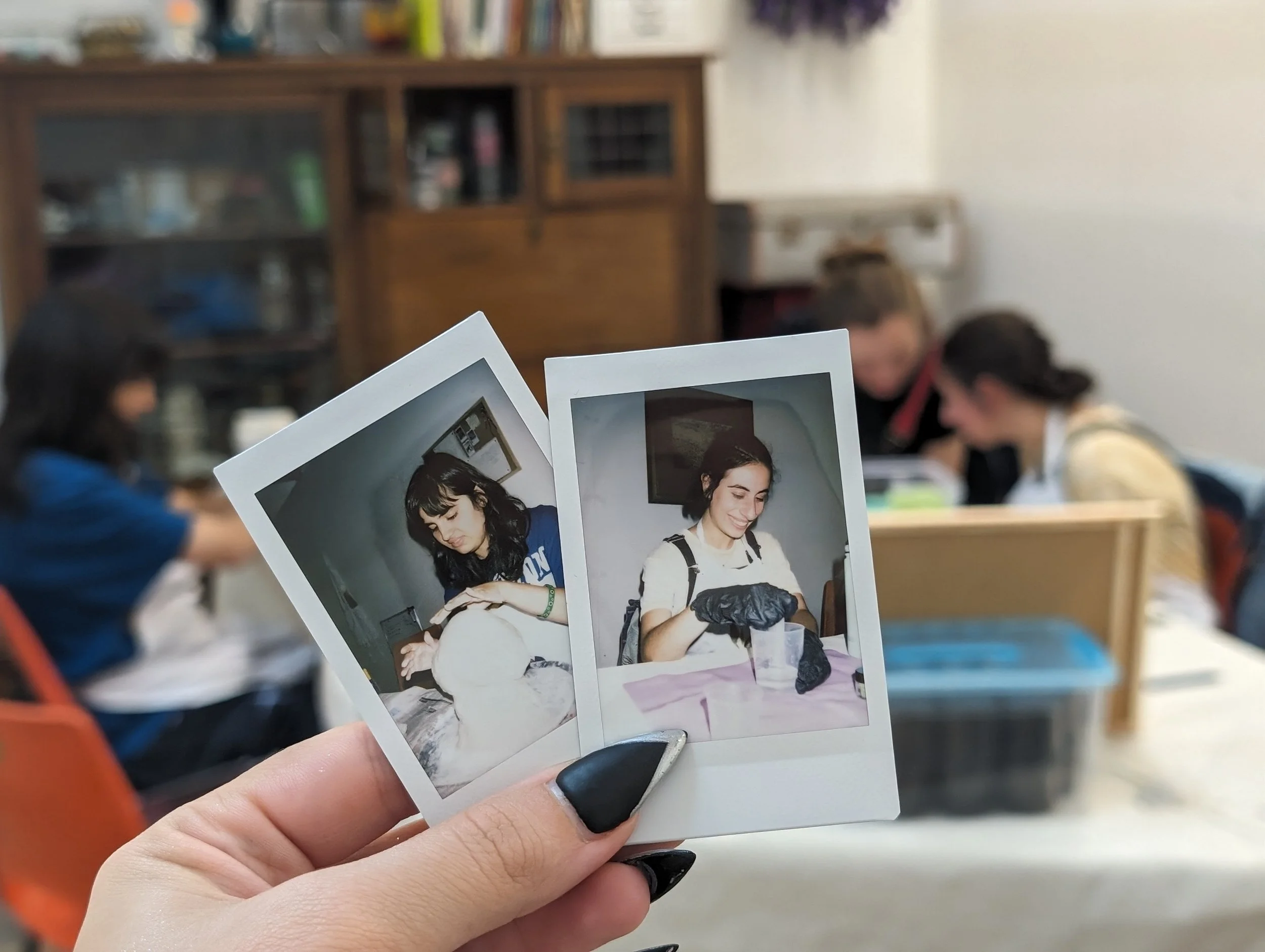Close-up of person holding printed photographs of a woman, with a classroom setting and students in the background.