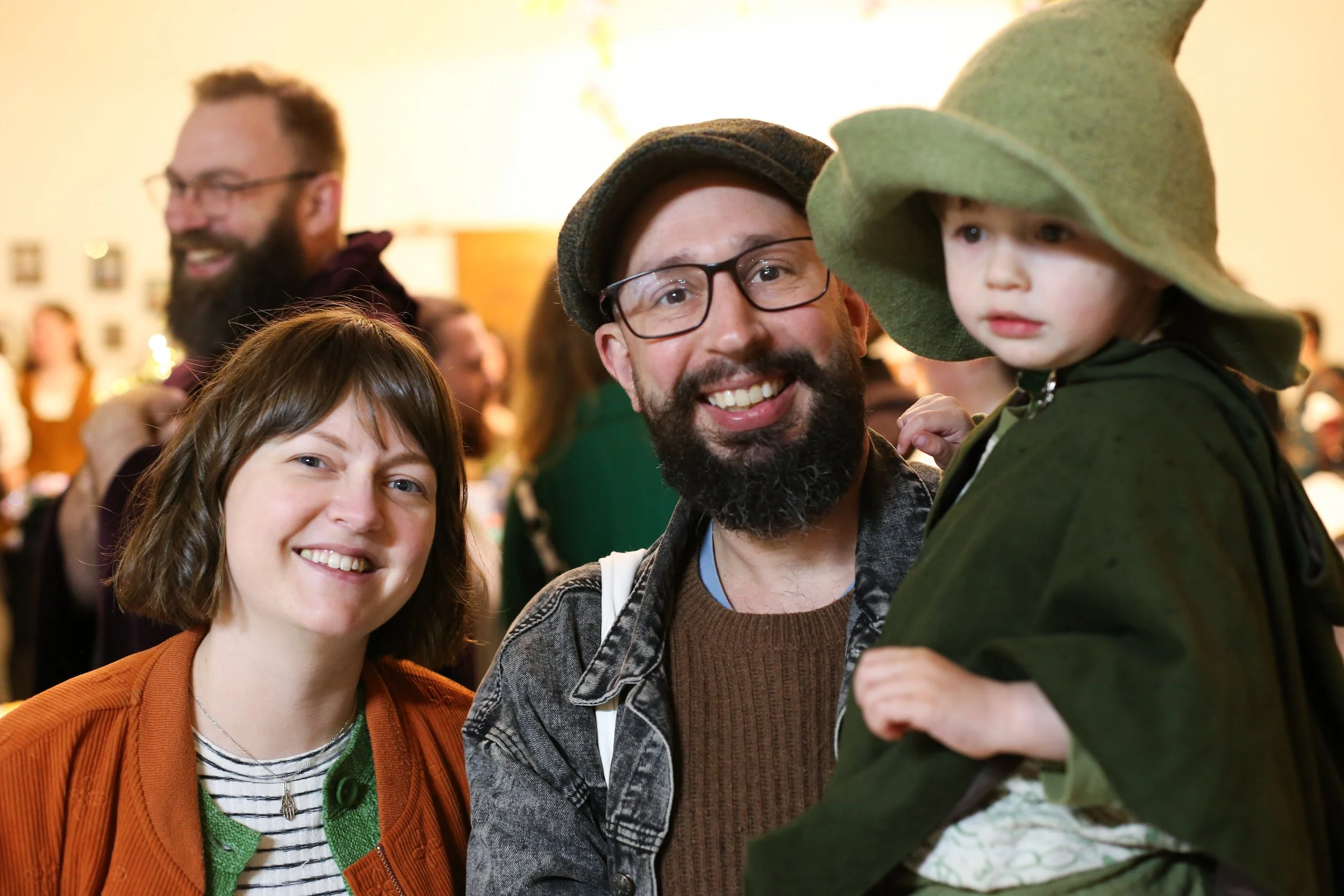 A group of smiling people at an indoor event, including a woman with short brown hair, a man with glasses and a beard wearing a hat, a young girl with a green coat and hat.