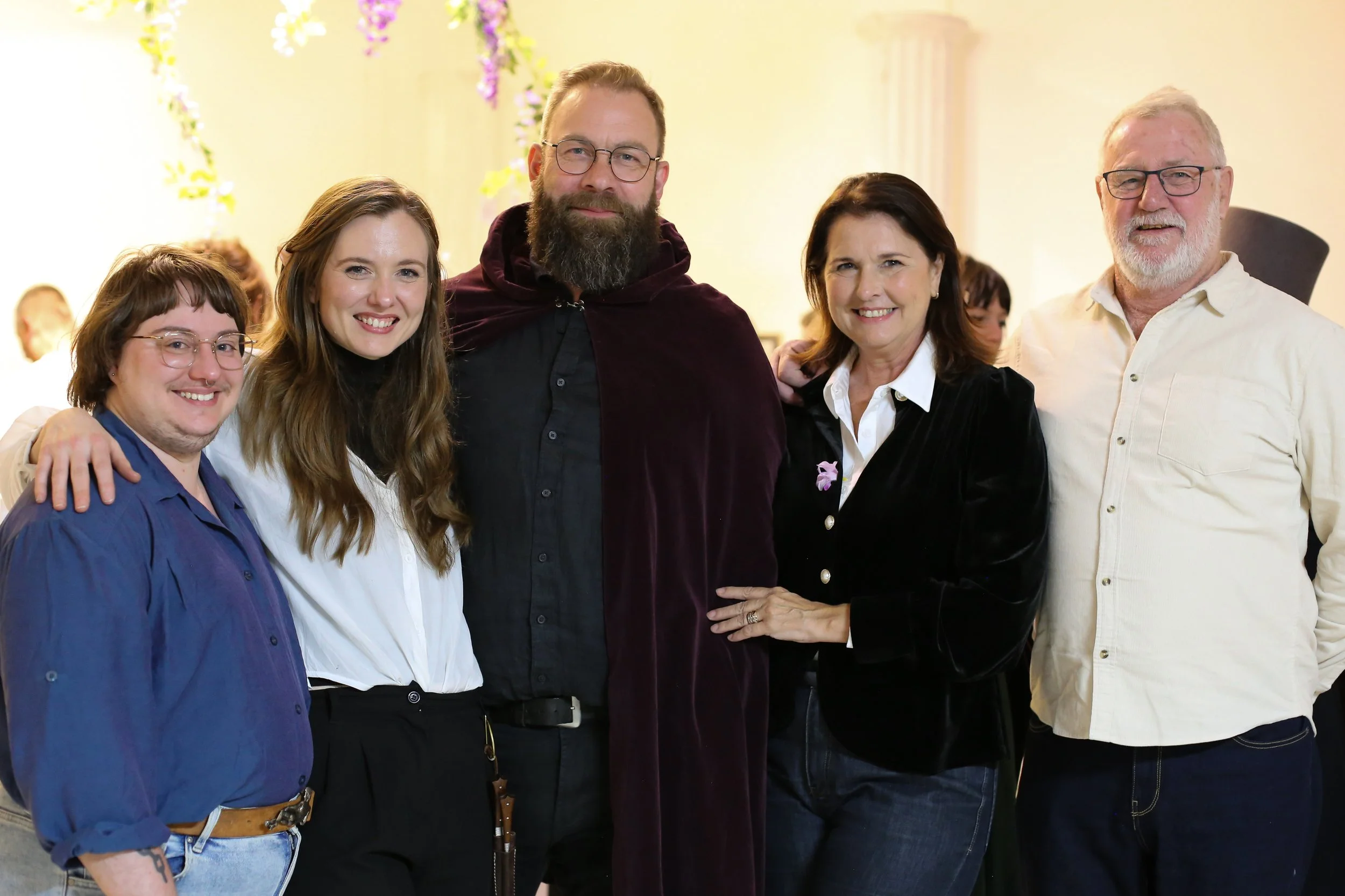 A group of five smiling adults standing close together indoors, with a blurred background featuring hanging purple flowers and a light-colored wall.