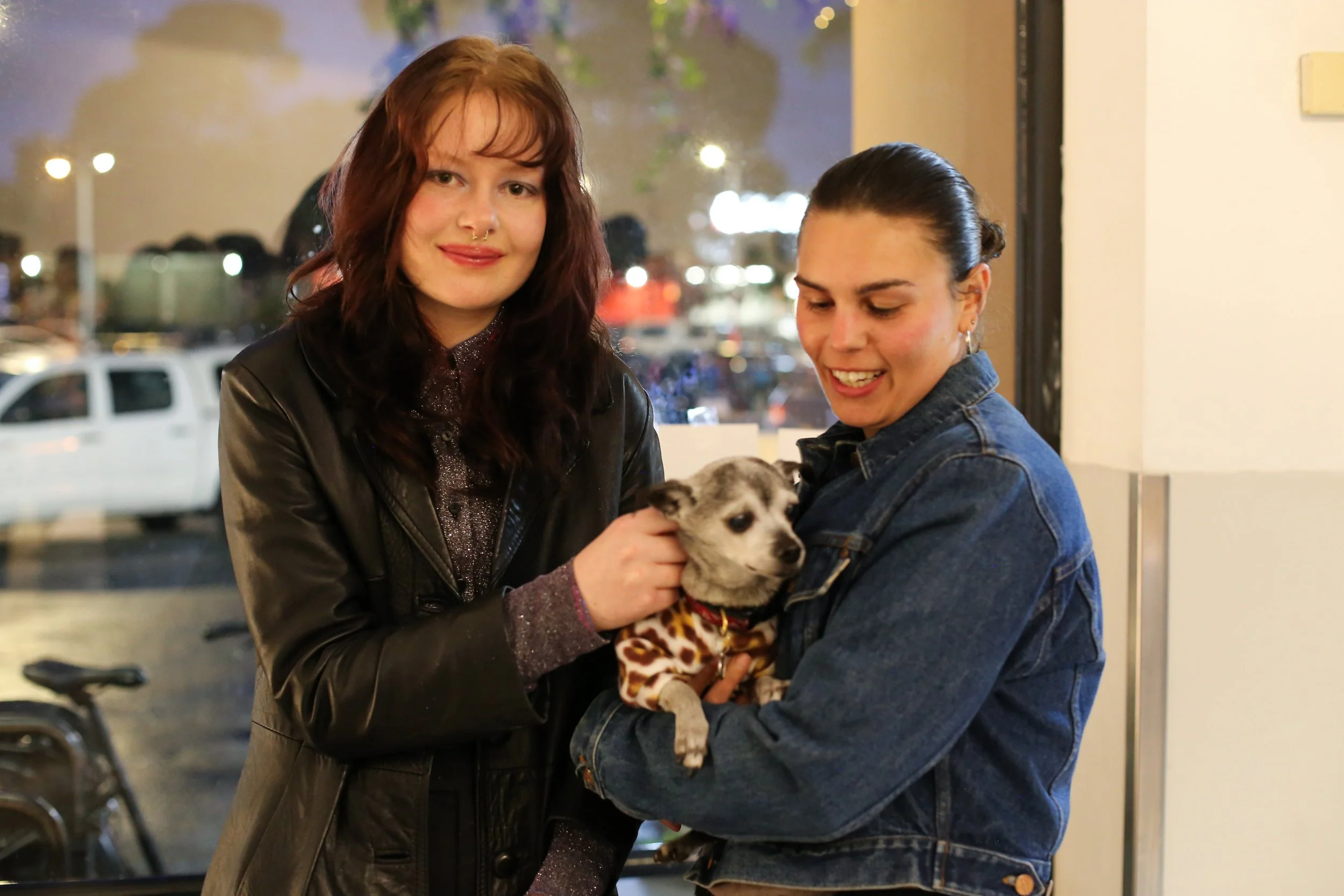 Two women holding a small puppy wearing a leopard print outfit inside a building with large windows, outside is a parking lot with cars and trees at night.