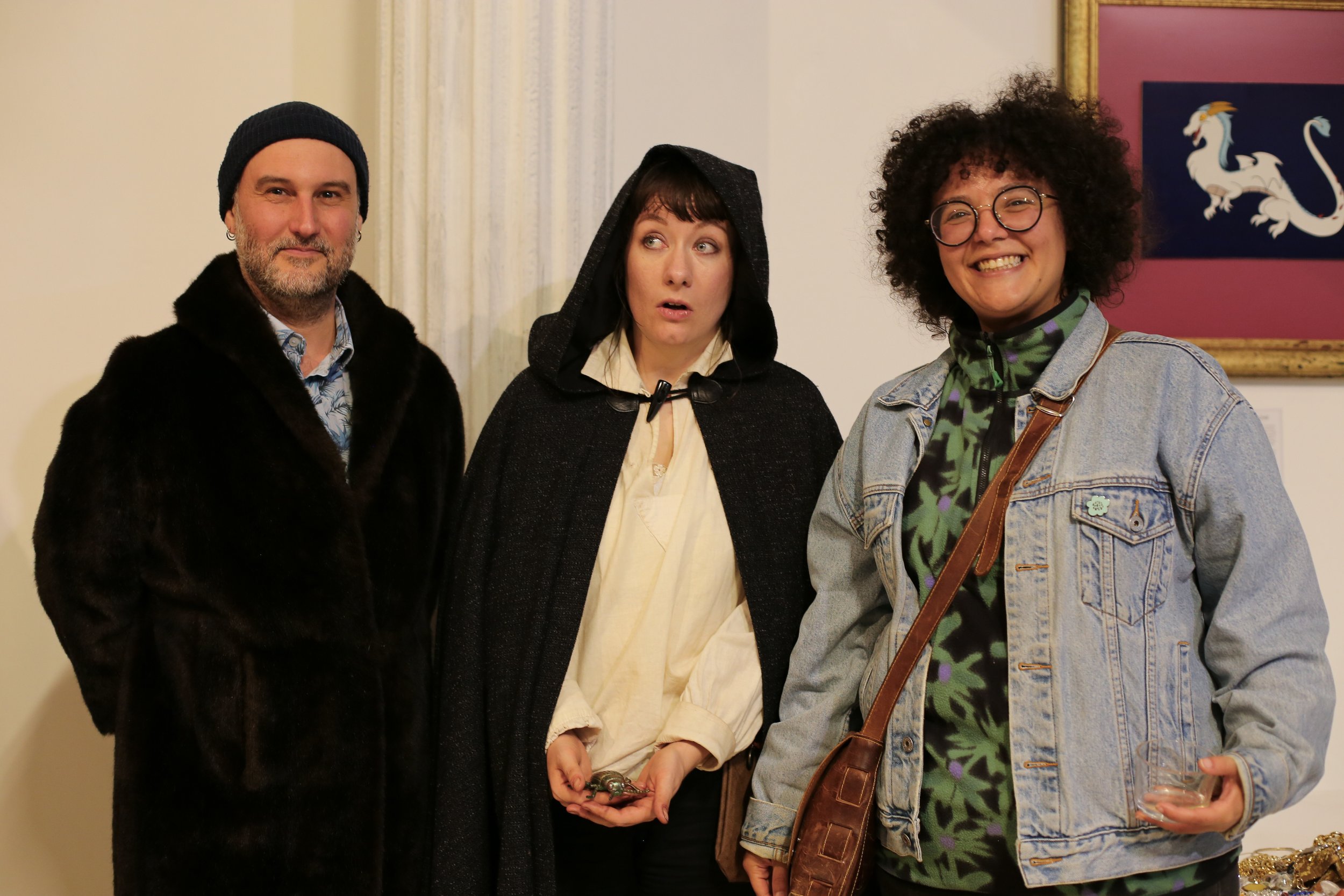 Three people standing indoors, posing for a photo. The person on the left is a man wearing a black fur coat and black beanie. The person in the middle is a woman with a black hooded cape, making a surprised face. The person on the right is a woman wi