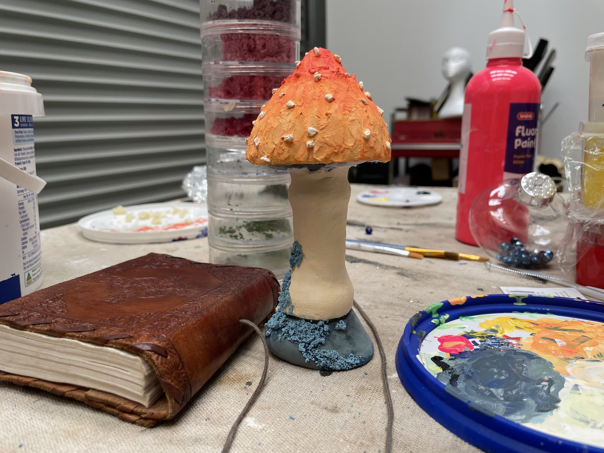 A small sculpture resembling a volcano with an orange top speckled with small white protrusions, a light-colored middle, and a darker base surrounded by blue textured material. The sculpture is on a wooden base and various art supplies, including pai