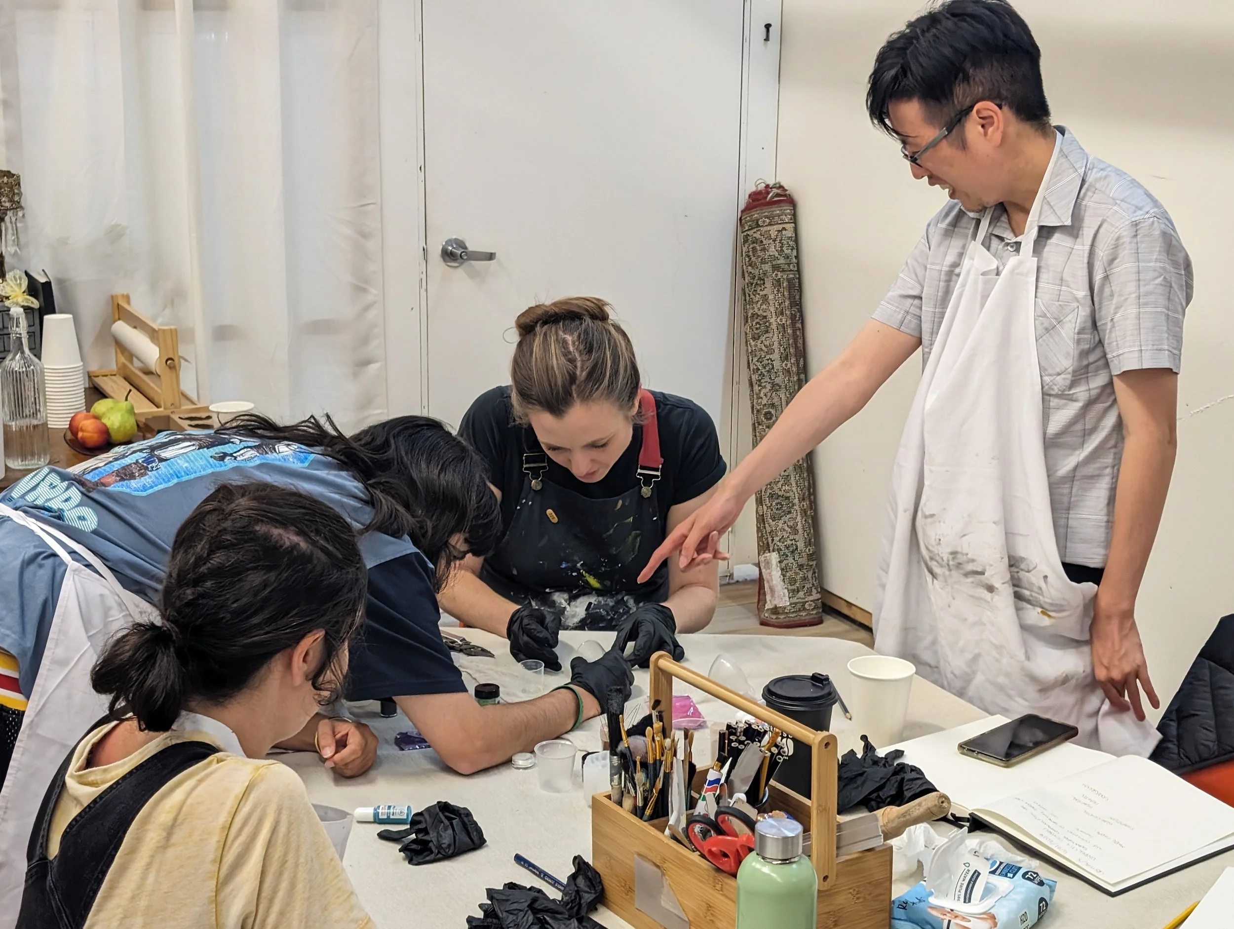 A group of people gathered around a table engaged in a hands-on activity or project, with tools and supplies such as scissors, glue, and brushes on the table, in a room with a plain white wall, with a man in glasses and an apron standing and pointing
