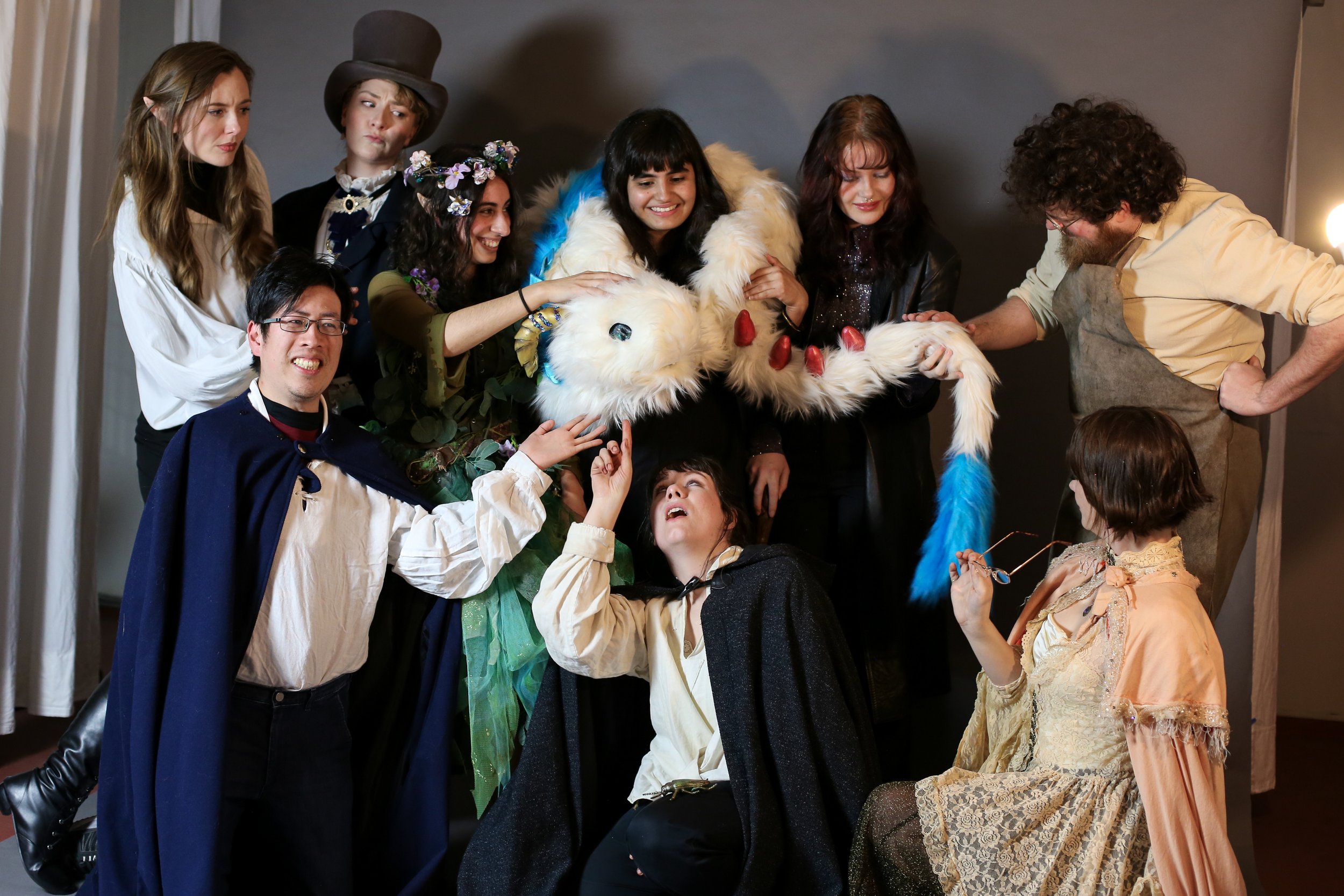 Group of people dressed in costumes, with some touching a large, white, furry creature with red horns and a blue tail, smiling and interacting in a room with gray walls.