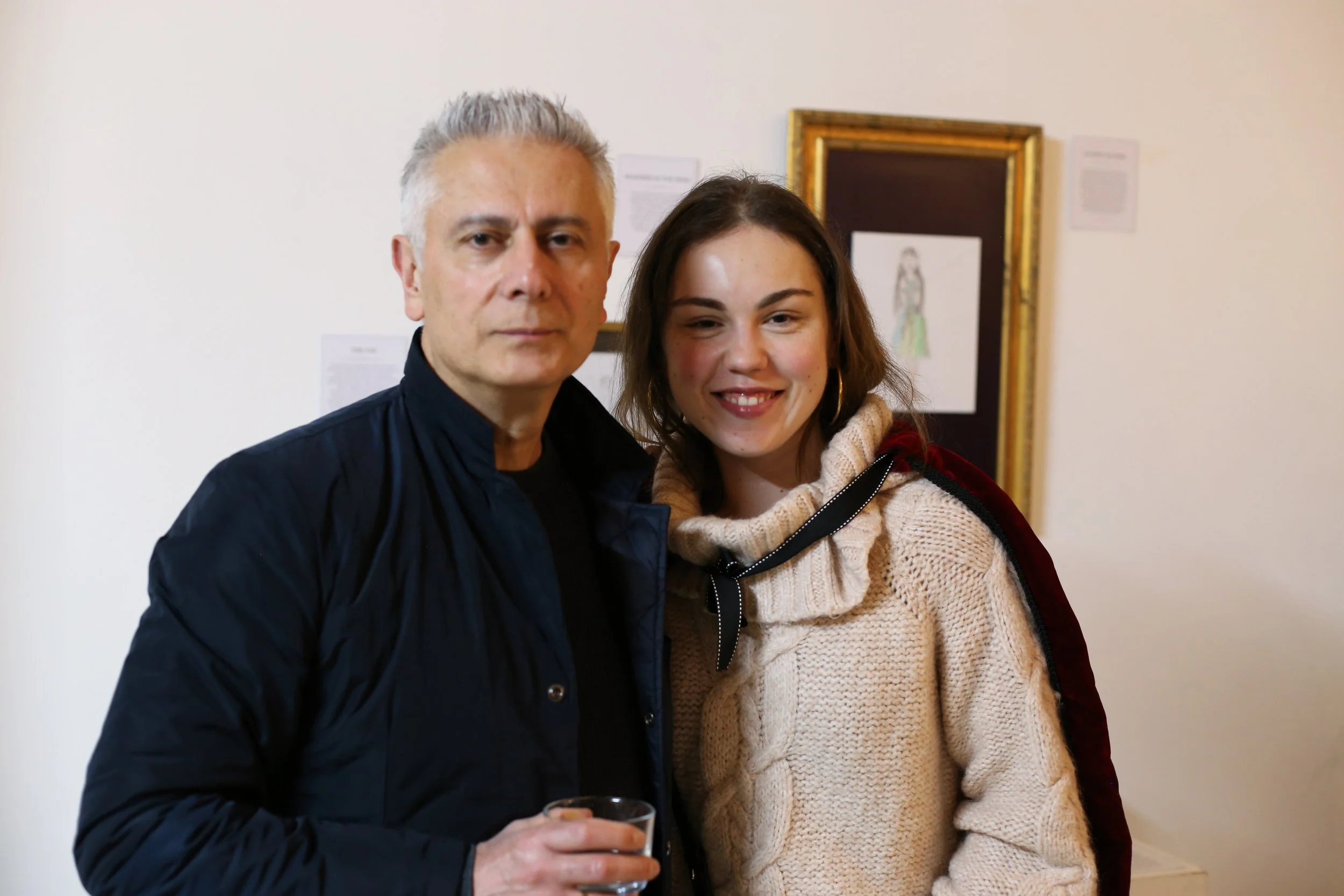 An older man with gray hair and a woman with shoulder-length brown hair standing close together indoors, smiling slightly, with artwork and papers on the wall behind them.