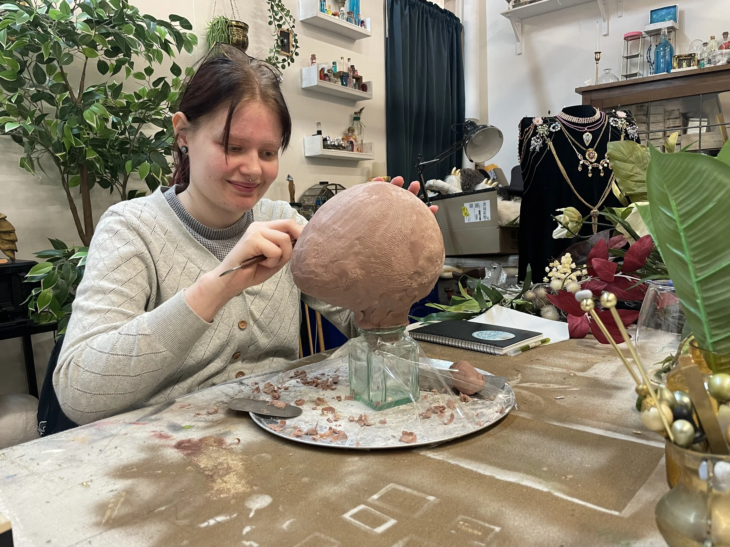 A woman with dark hair and a septum piercing is sculpting a large, rounded clay sculpture at a table. The workspace is decorated with various plants, flowers, and jewelry displays.