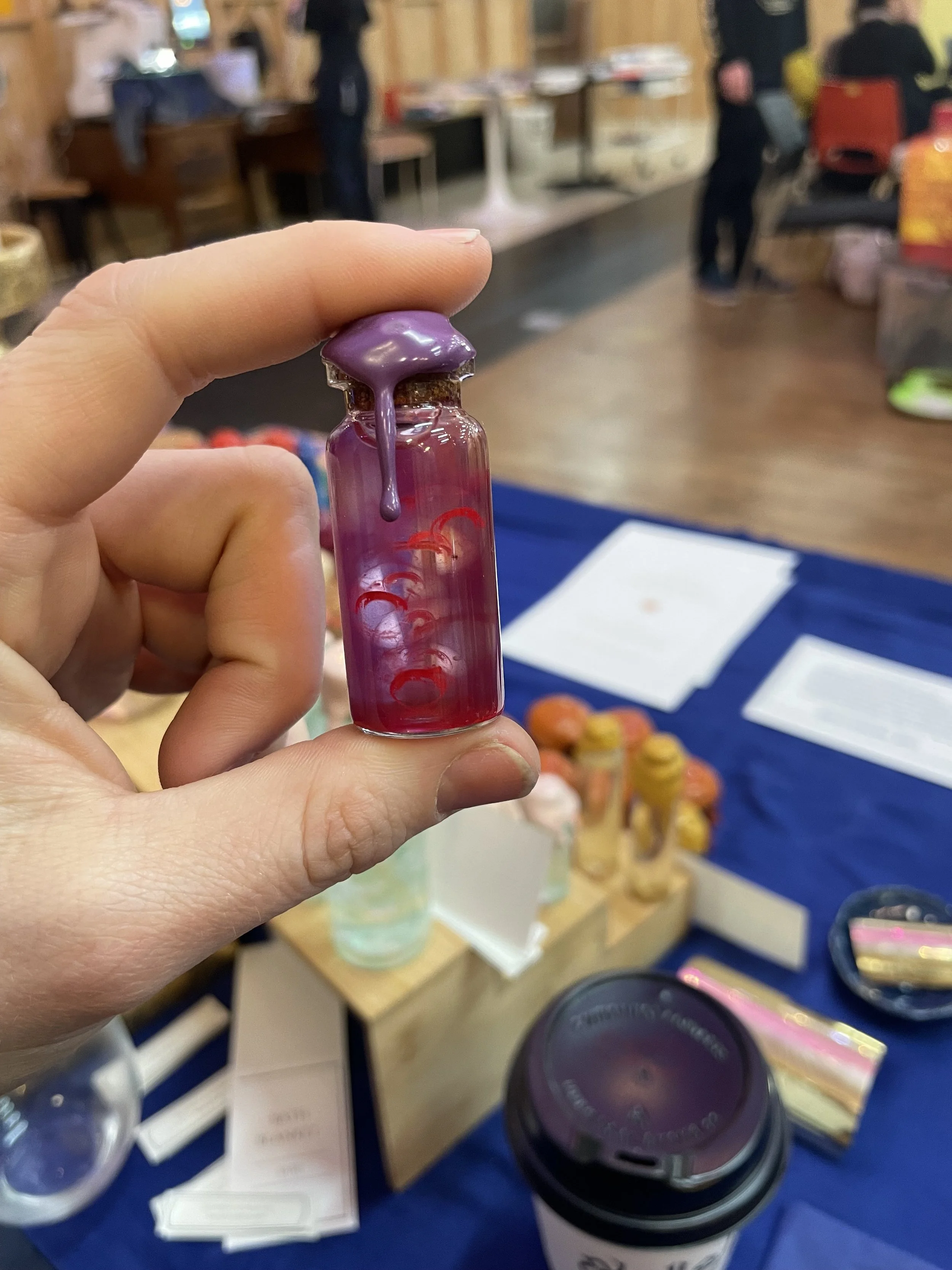 A hand holding a small glass vial with purple liquid dripping from the top, at an indoor booth or store with various jars and containers on tables in the background.