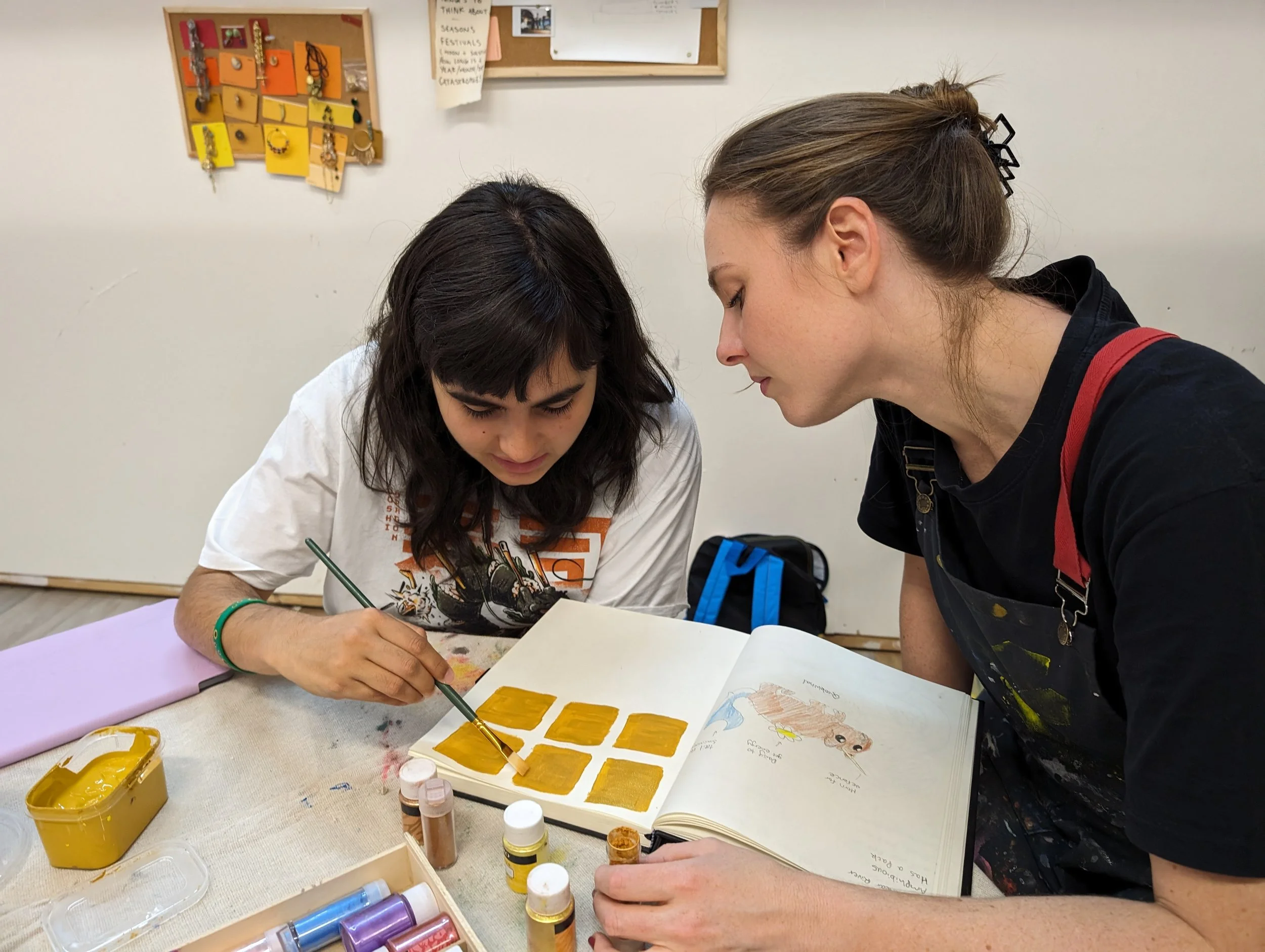 Two women are working together on an art project at a table; one is painting golden rectangles on paper, while the other looks on closely. art supplies and notebooks are scattered on the table, and a bulletin board with notes and jewelry hangs on the