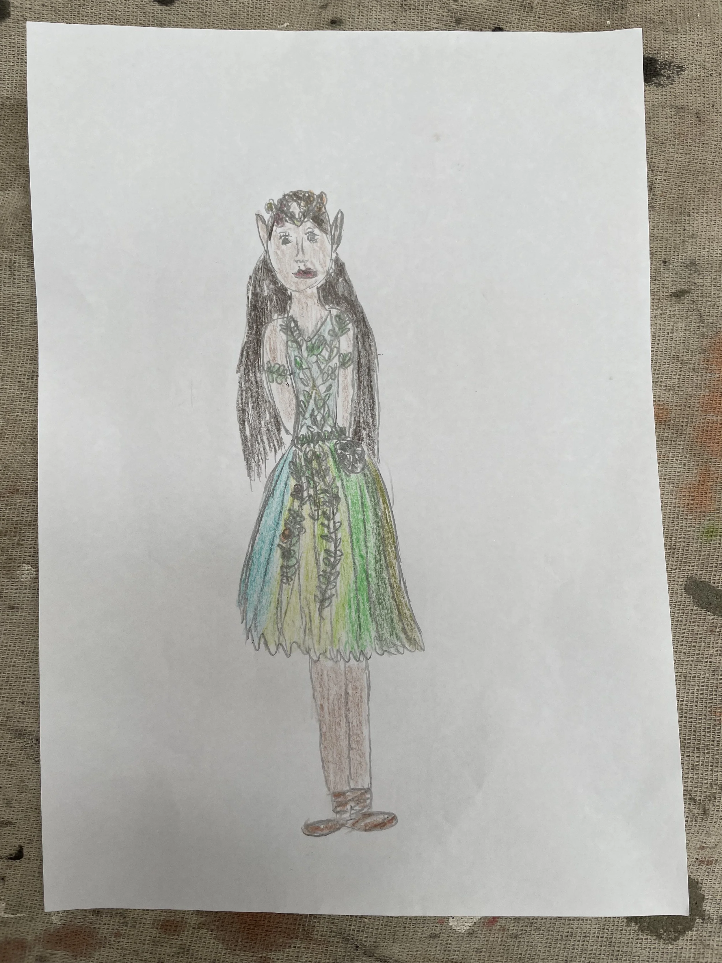 A child's drawing of a fairy or elf with pointed ears, long dark hair, wearing a green and yellow dress with leaf patterns, and standing on bare feet.