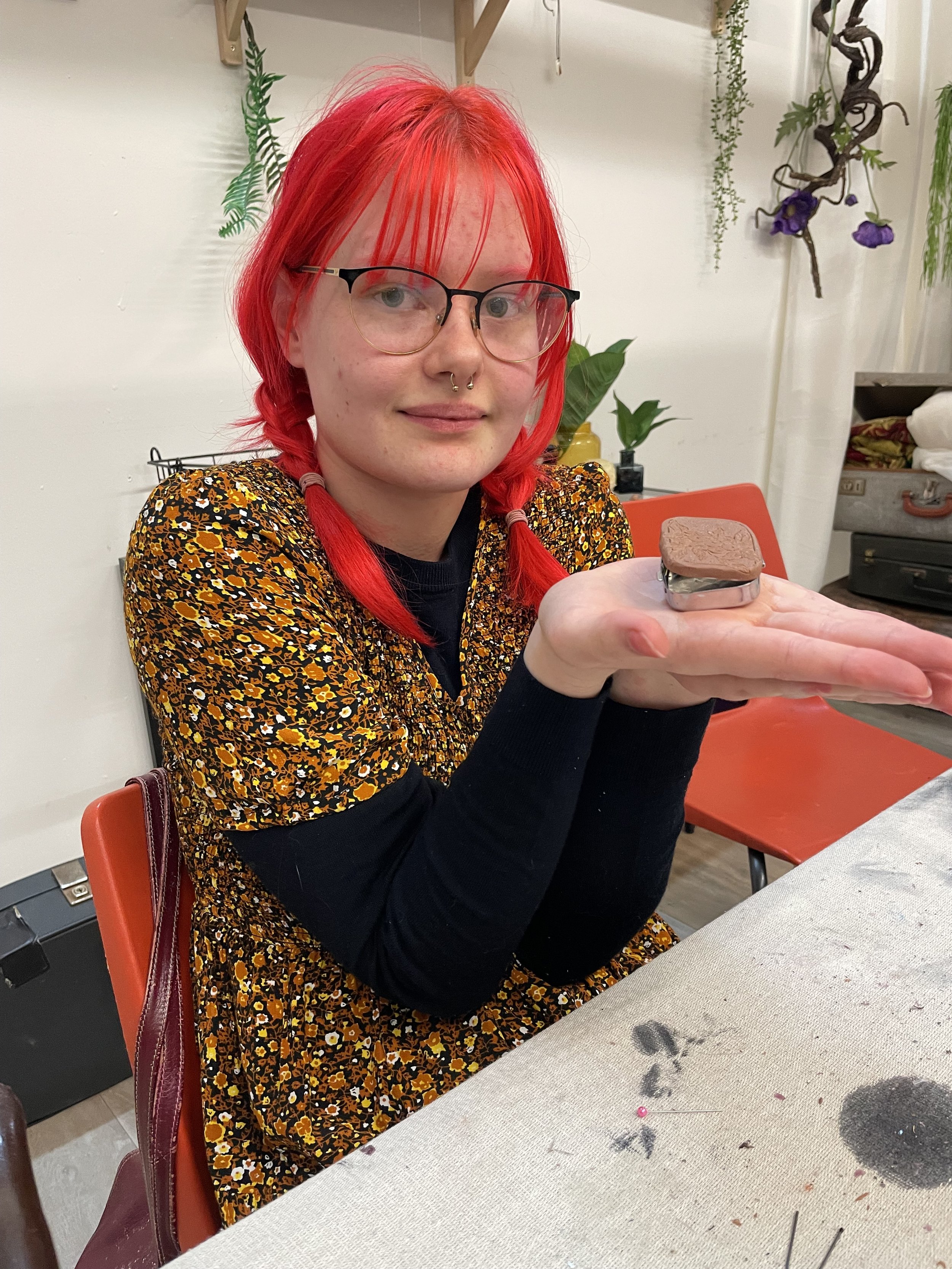 A young person with bright red hair, glasses, and a nose ring, sitting at a table and presenting a small, square, chocolate-colored box with a textured surface.