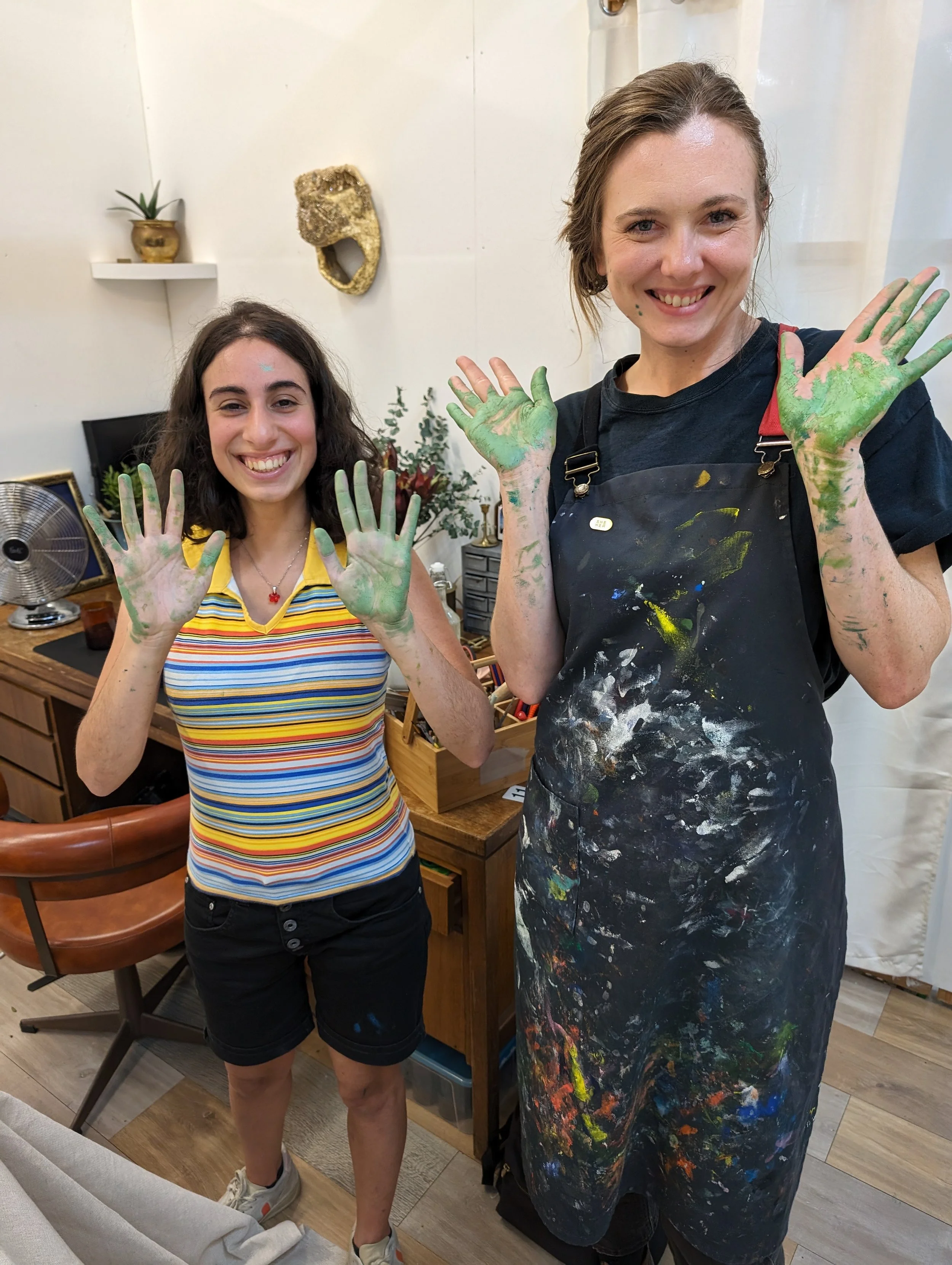 Two women smiling and showing their hands covered in green paint. One woman has curly dark hair, wearing a striped shirt, and black shorts. The other woman has short wavy hair, wearing a black apron with paint splatters.
