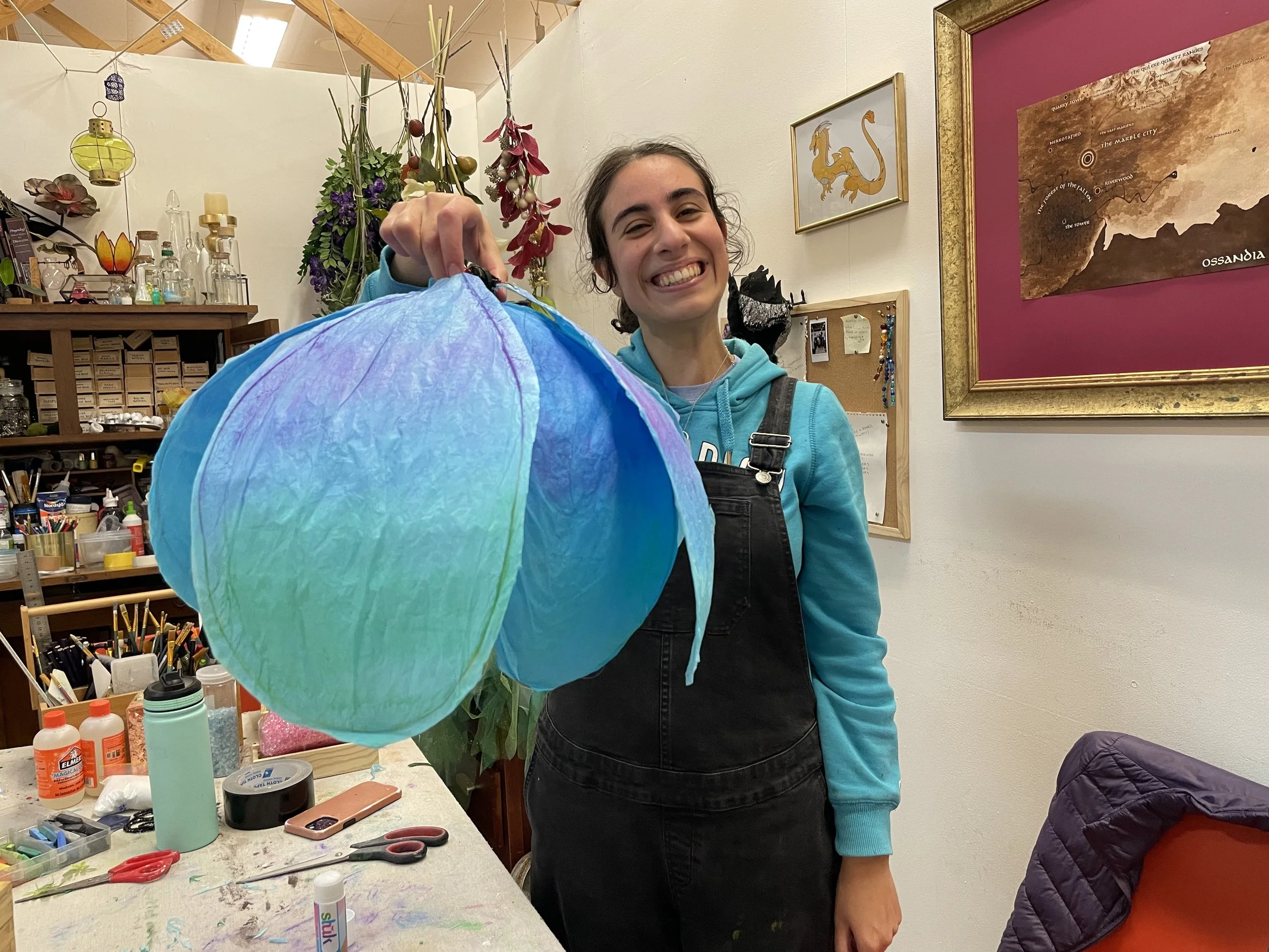 Smiling woman holding colorful paper lantern in art studio with craft supplies, framed artwork, and plants in background.