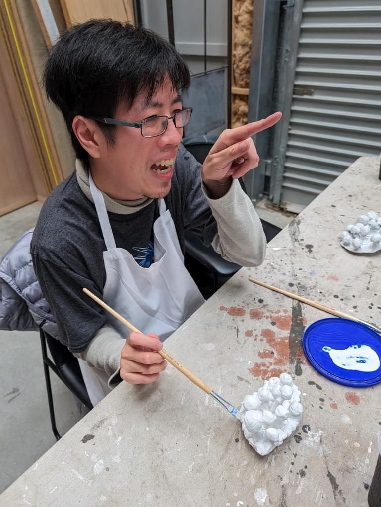 A man wearing glasses and an apron is smiling and gesturing while working on a craft project with white plaster or clay, using a paintbrush. There are additional craft materials on the table.