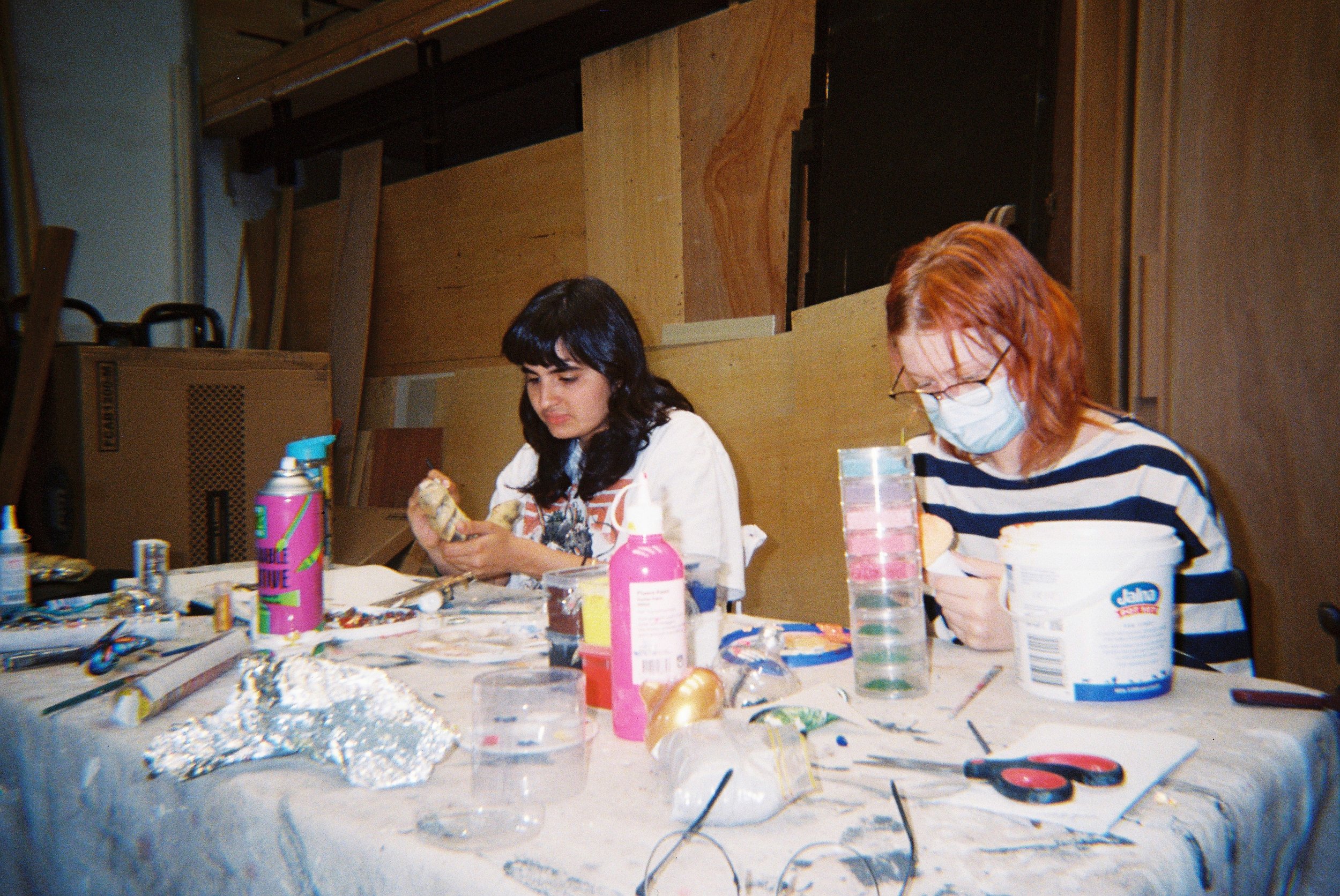 Two women sit at a cluttered table working on arts and crafts projects. The table is filled with various craft supplies, including glitter, scissors, and bottles of paint. One woman has dark hair and is wearing a white shirt, while the other has red 
