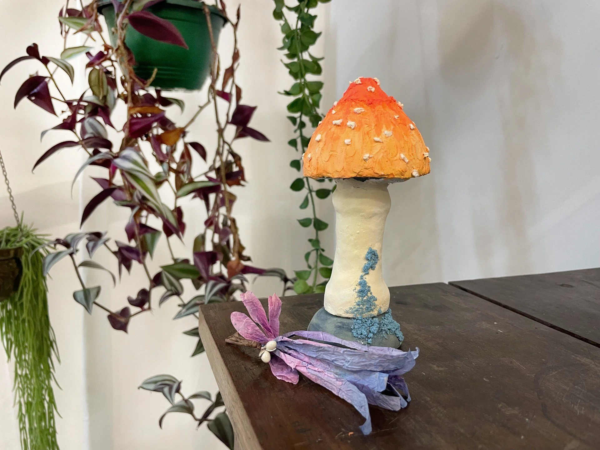 Decorative mushroom sculpture with a beige stem and orange cap, placed on a dark wooden table with purple and pink wrapped leaves and white berries, surrounded by hanging and potted green plants.