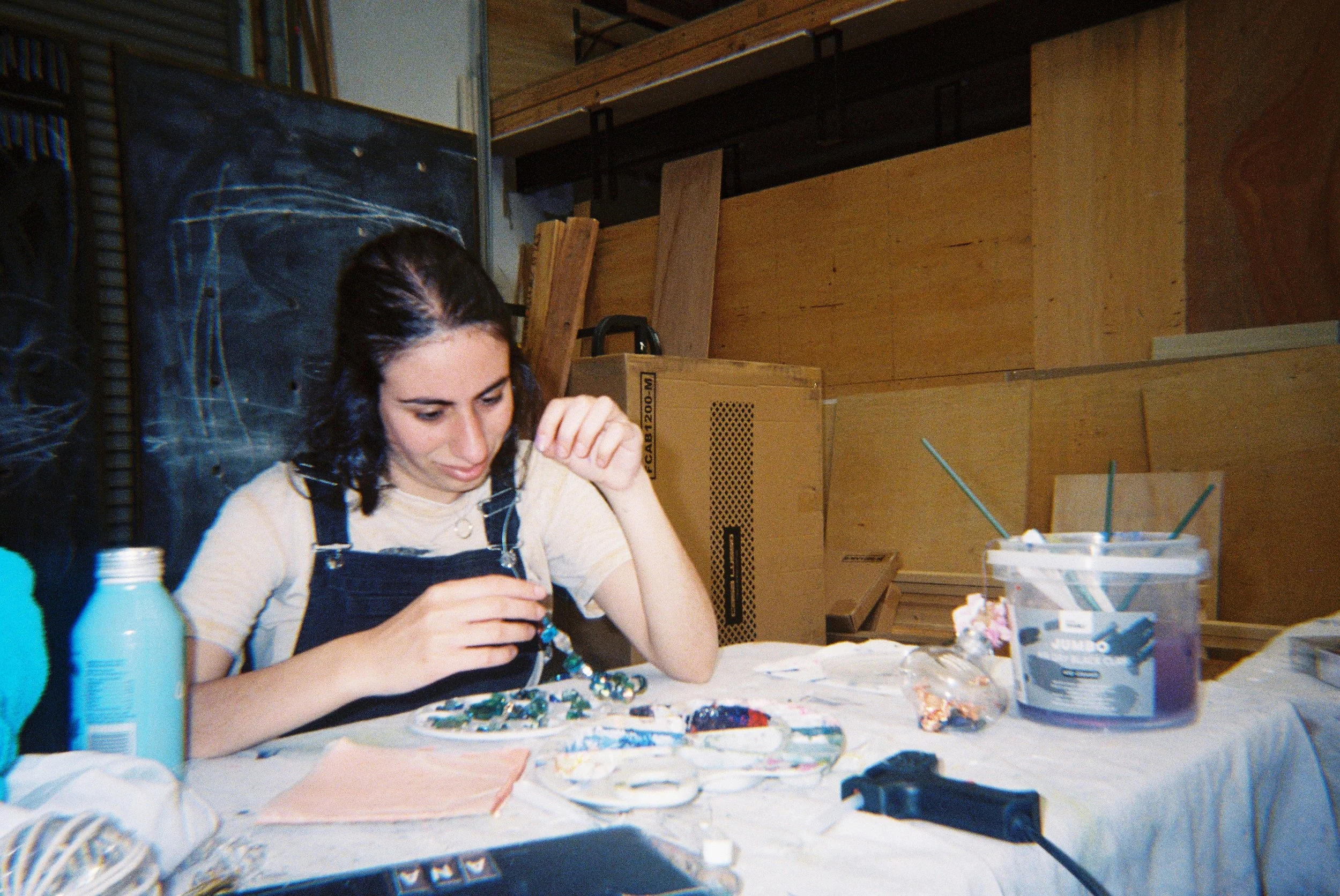 A woman with dark hair wearing a striped shirt and overalls is sitting at a cluttered table in a workshop or craft room, working on a craft project. The table has various craft supplies and tools, including bottles, paint, and fabric. In the backgrou