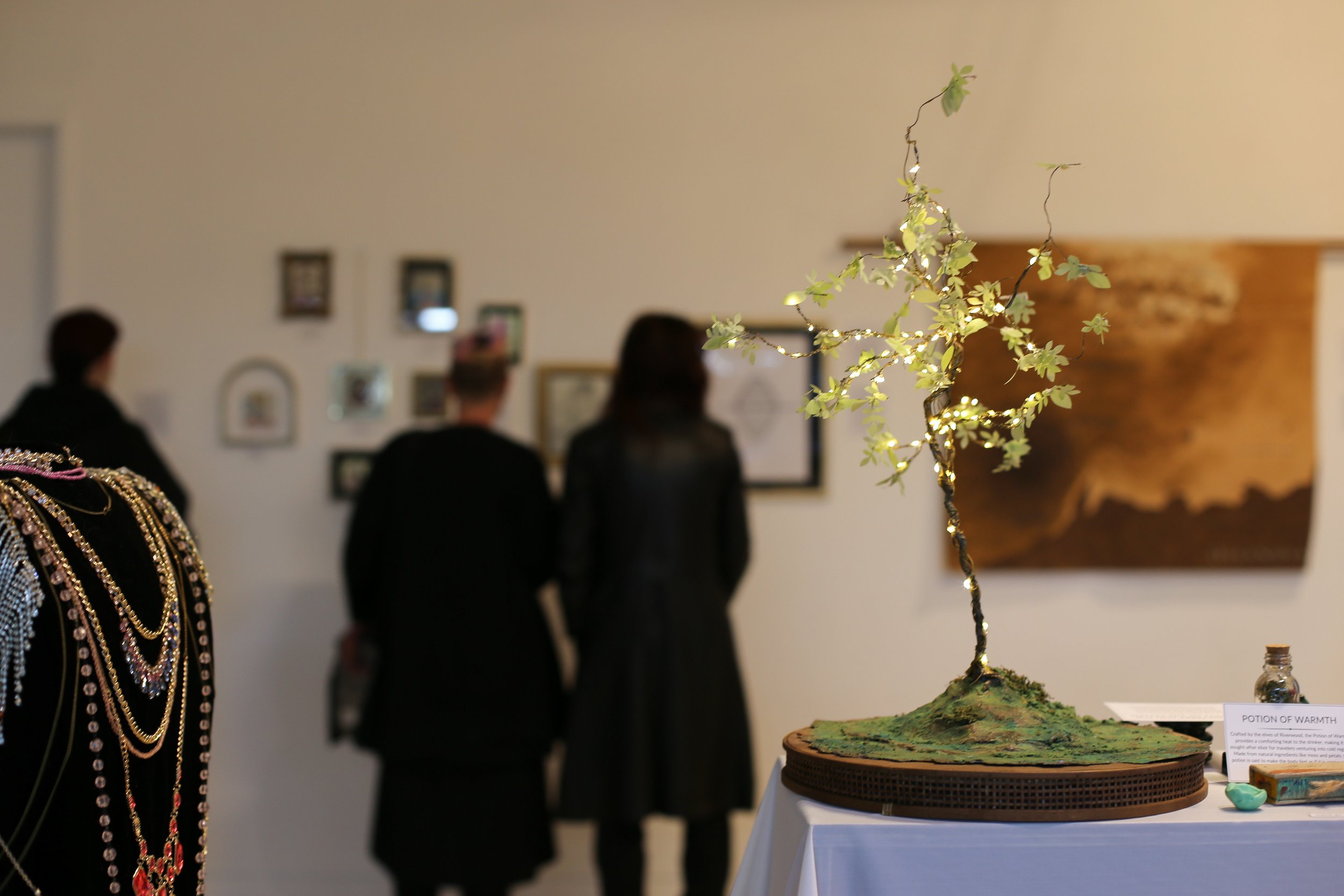 A decorative tree with lights on it on display at an art exhibit, with blurred figures of people looking at artwork in the background.