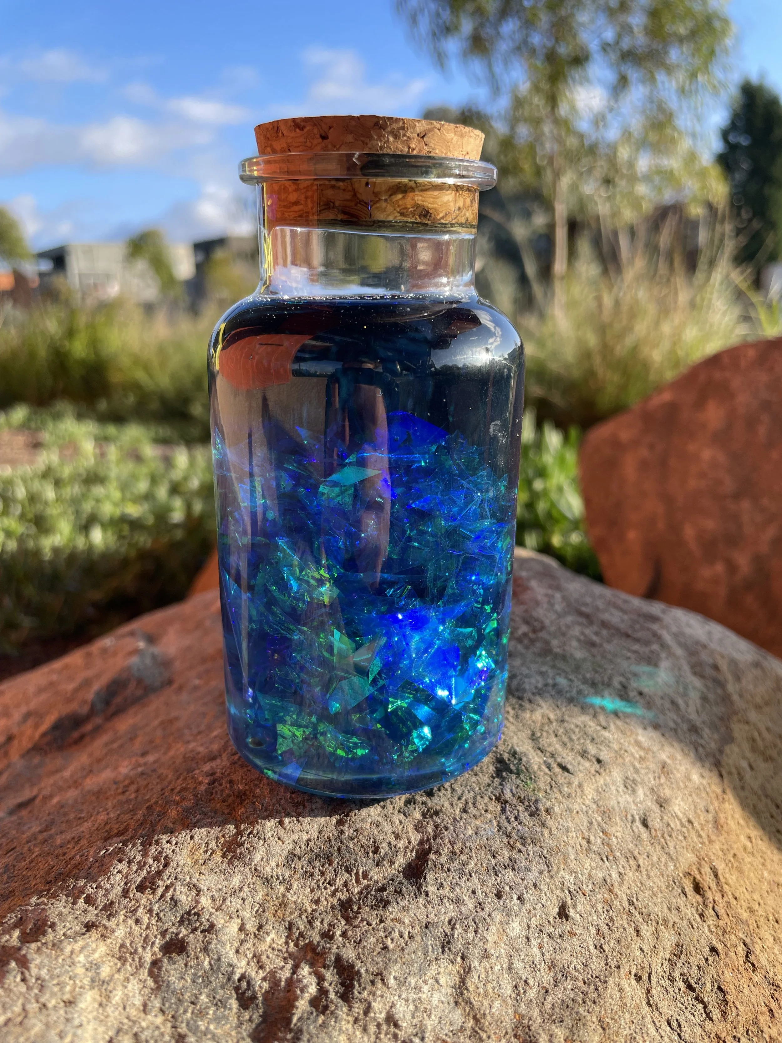 A glass jar filled with blue decorative glass fragments, sealed with a cork lid, sitting on a large rock outdoors. The background features greenery, trees, blue sky with clouds, and some distant buildings.